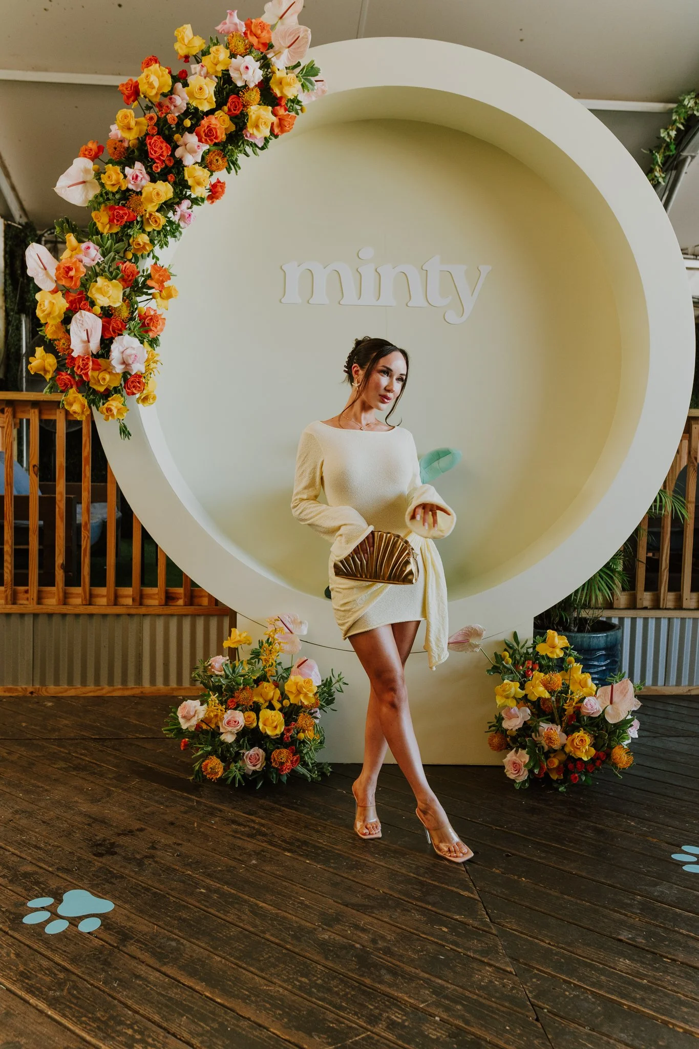 A woman with dark hair styled in an updo, wearing a cream-colored dress, clear high heels, and holding a fan, stands in front of a large white circular backdrop decorated with colorful flowers with the word "minty" displayed behind her.