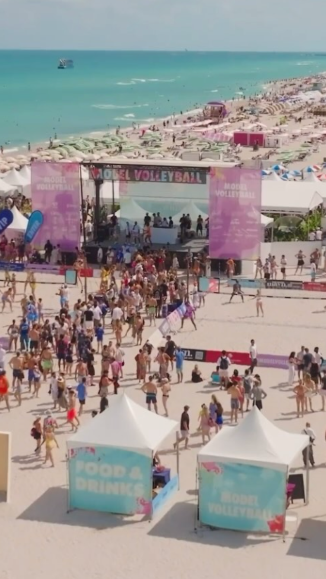 Beach scene with many people, umbrellas, and a volleyball event stage with banners reading "Modèle Volleyball." The ocean is in the background.