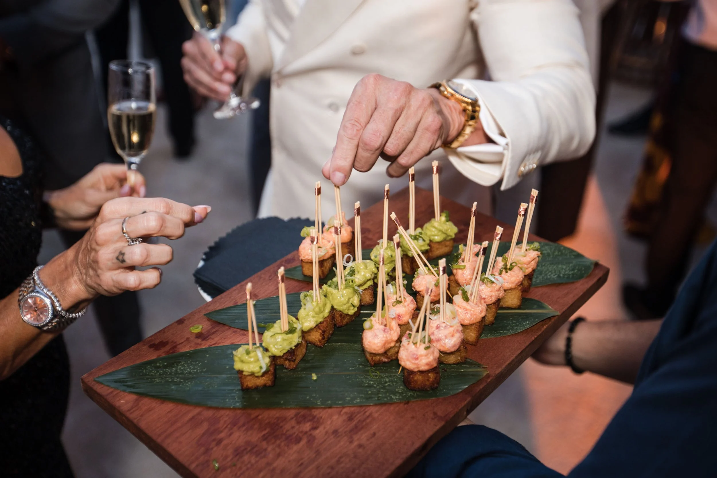 People at a celebration serving and sharing appetizers topped with green and pink garnishes on a wooden tray with leaves. One person is wearing a gold watch and bracelet.