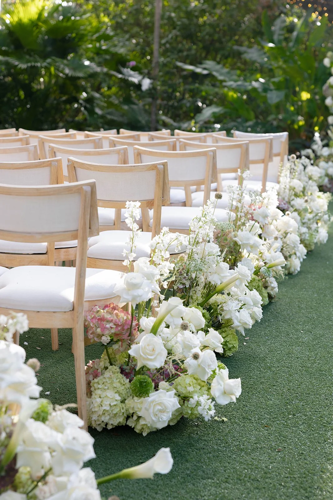 Outdoor wedding ceremony setup with empty wooden chairs and white floral arrangements along the aisle, on a grassy surface surrounded by greenery.