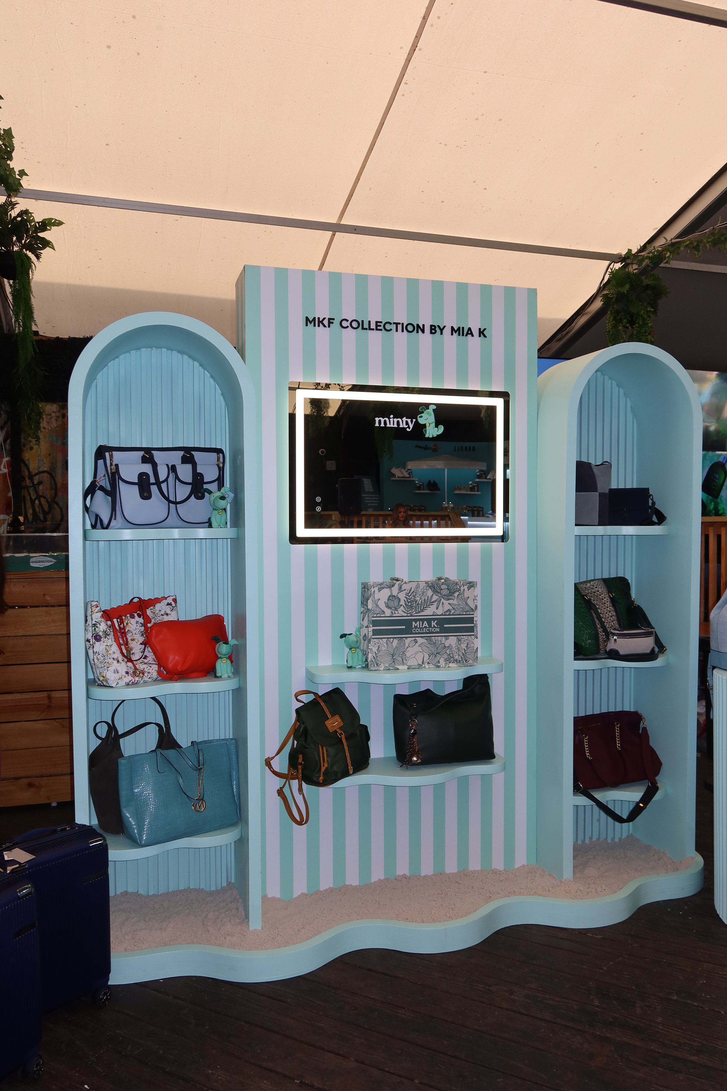 Display stand with handbags and accessories, branded as 'MKF COLLECTION BY MIA K' with pastel blue and pink striped design.