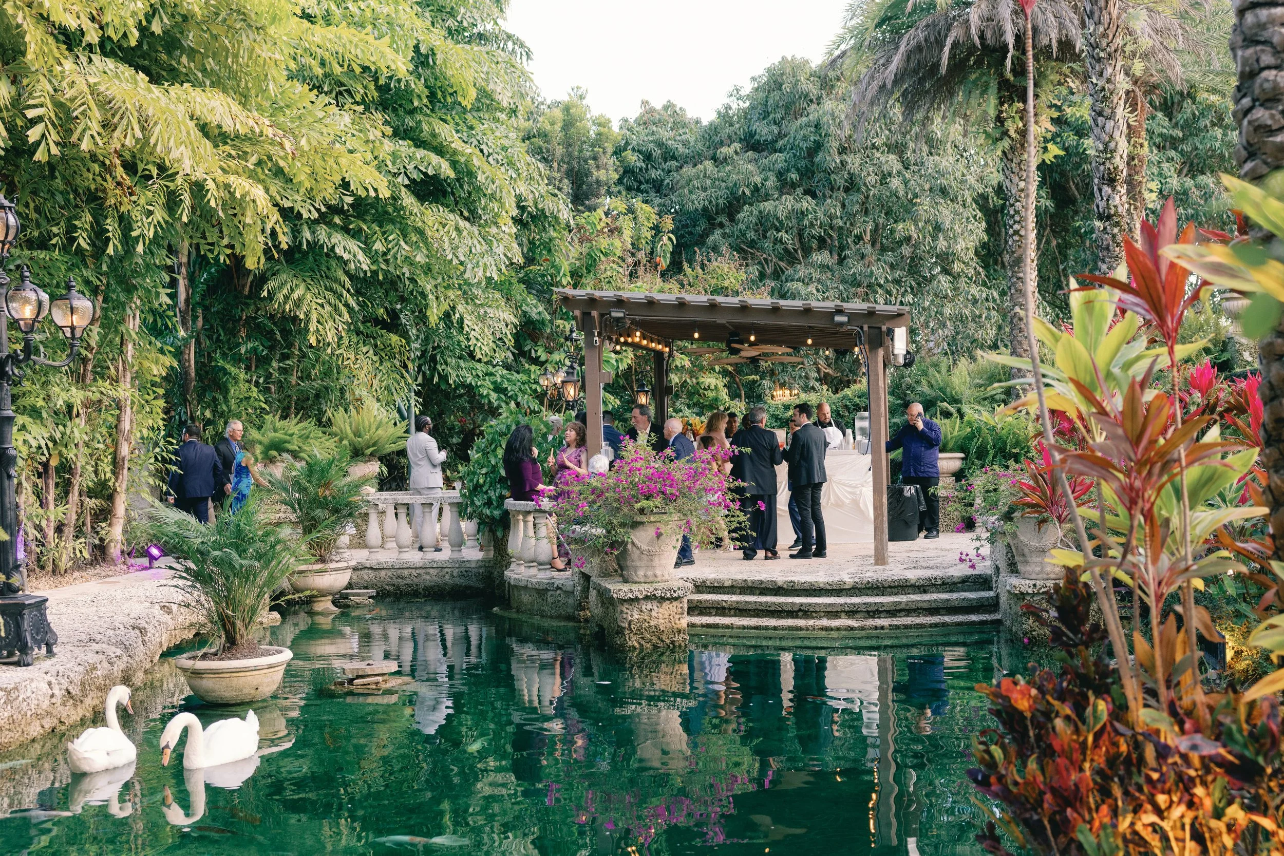 People gather under a wooden pavilion at a garden party surrounded by lush greenery, including tall trees and colorful flowering plants, with swans swimming in a pond in the foreground.