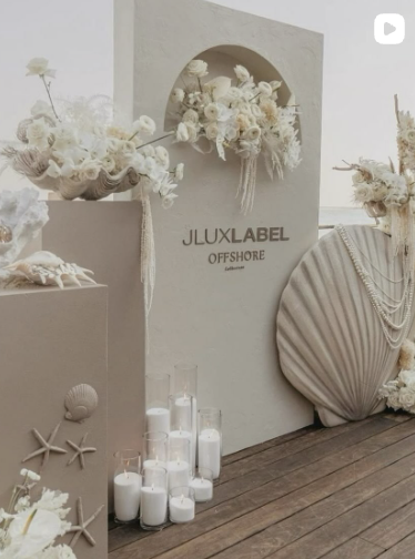 Decorative display with white flowers, candles, seashells, starfish, and a large fan-shaped object, featuring a sign that says 'JLUXLABEL OFFSHORE'.