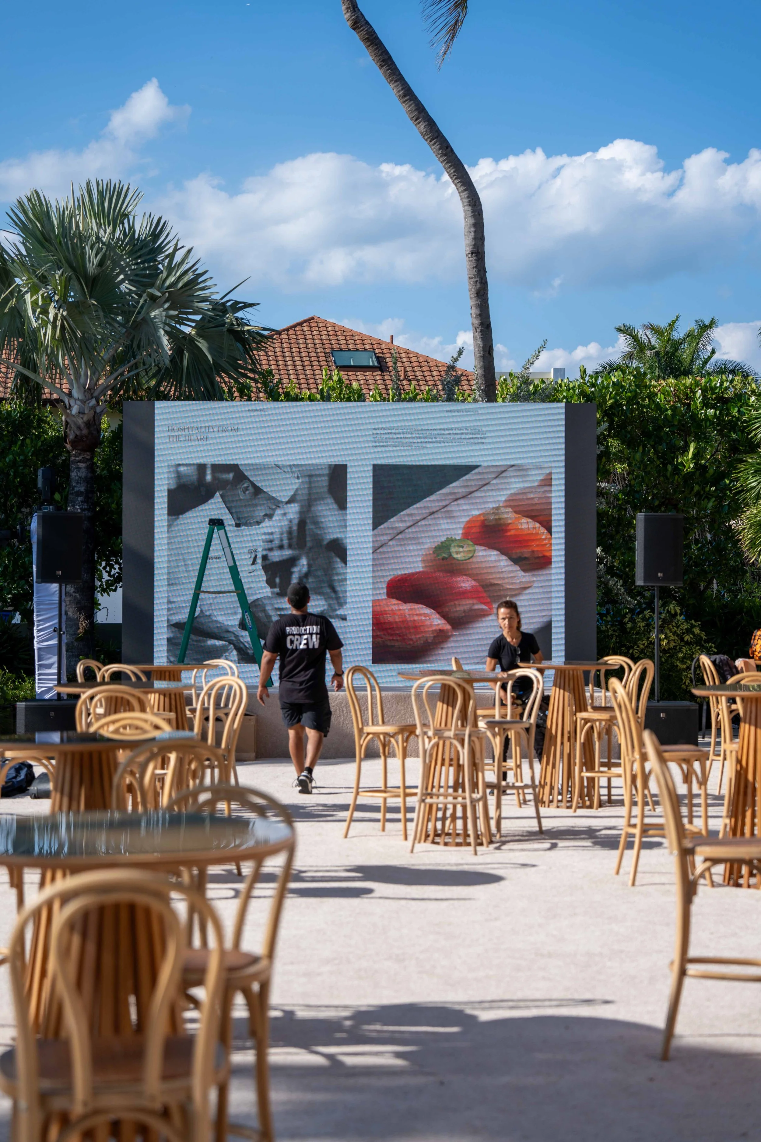 An outdoor scene with empty wooden chairs arranged in a courtyard, a large digital screen displaying abstract artwork, people setting up equipment, tropical trees, and a blue sky with clouds.