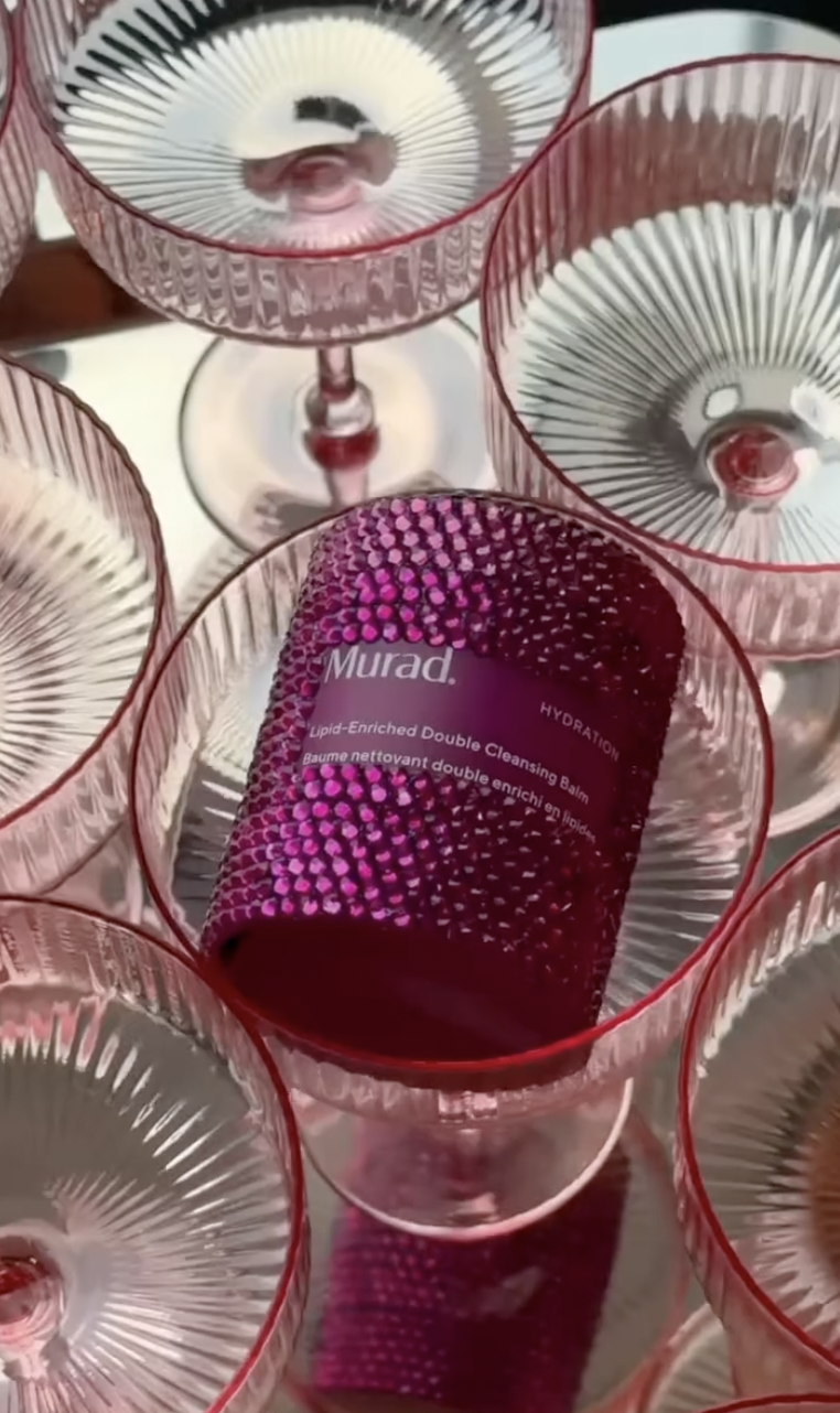A pink cylindrical bottle of Murad Lipid-Enriched Double Cleansing Balm surrounded by multiple empty pink and clear glass champagne glasses.