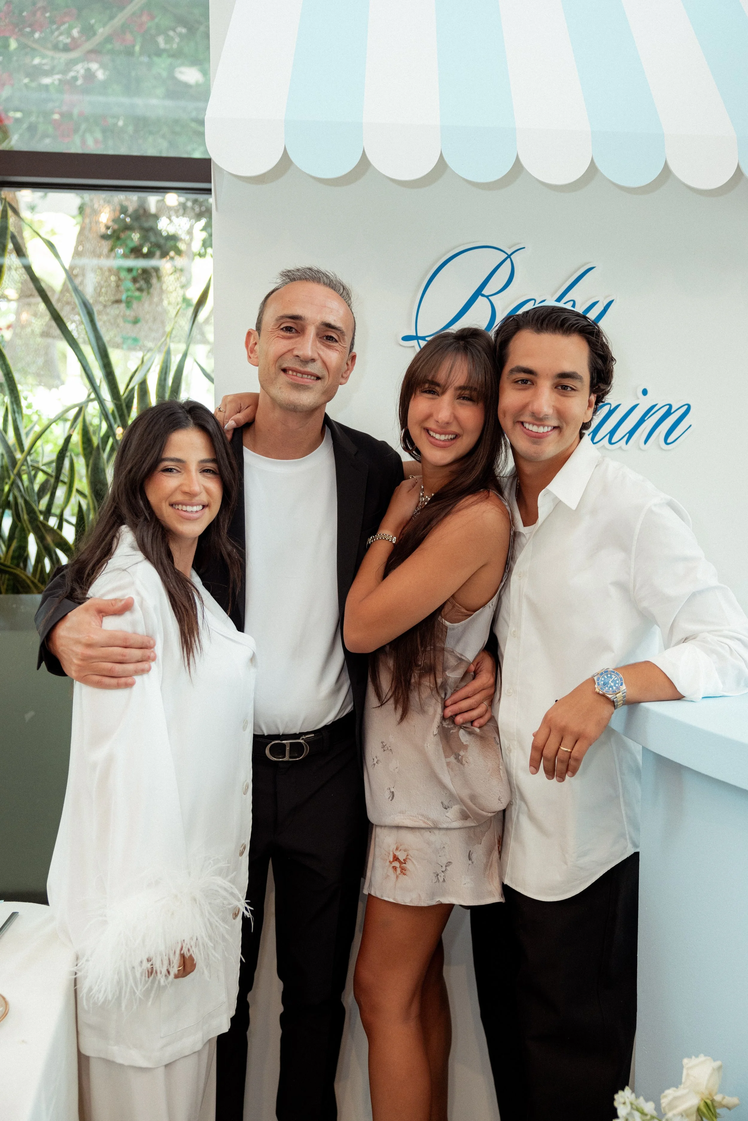 Group of four people smiling and posing together at an event, standing in front of a white wall with part of the word 'Beauty' visible, with windows and greenery in the background.