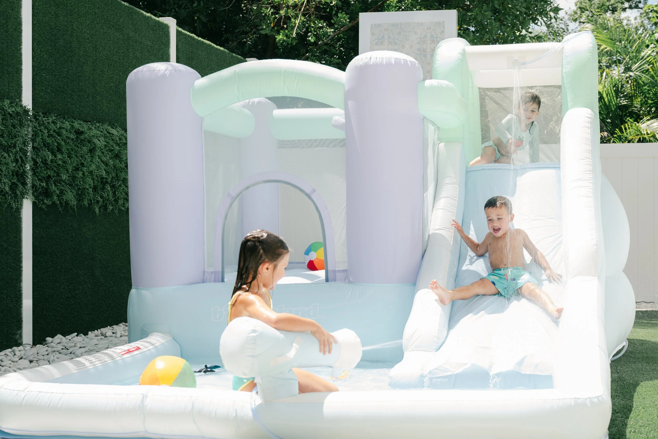 Children playing on an inflatable water slide in a backyard, with some children in the pool and others climbing the slide, surrounded by green foliage.