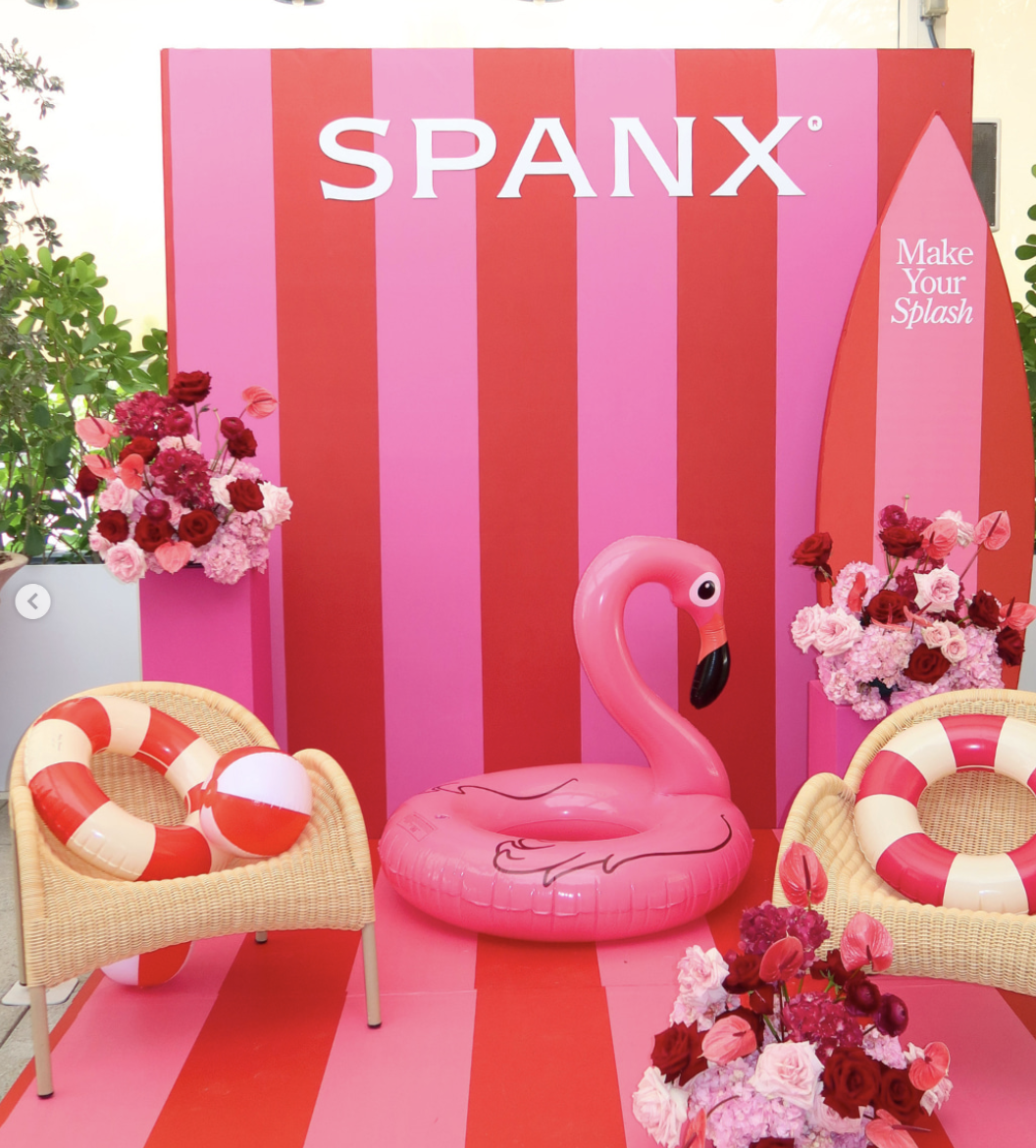 A pink striped backdrop with the brand name SPANX, surrounded by pink and red floral arrangements, a pink flamingo pool float, two wicker chairs with red and white life preservers, and pink and red items indicating a summer or pool party setting.
