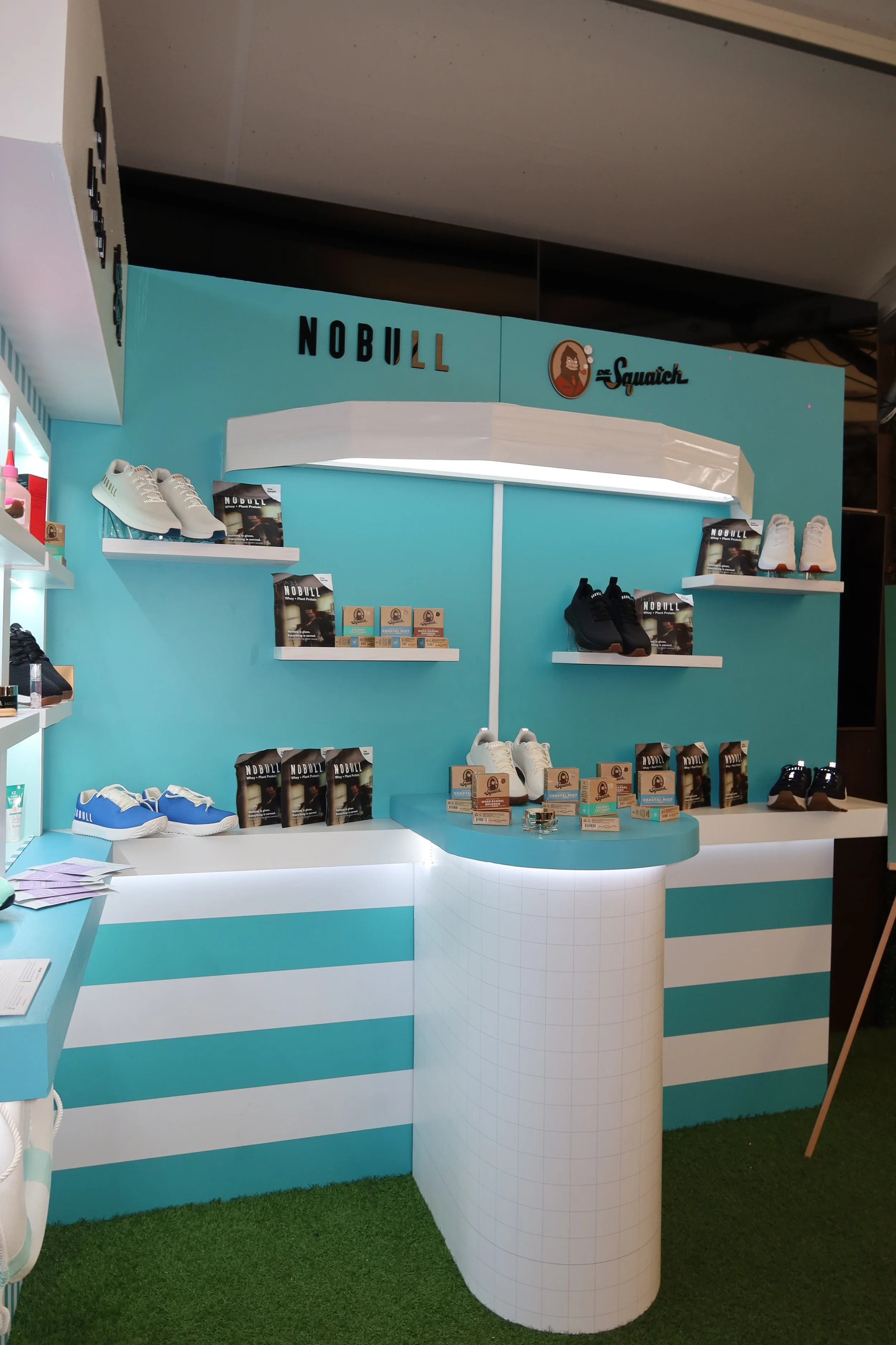 Store display with sneakers on shelves and promotional materials, featuring the brand Nobull and Dr. Squatch, with a light blue and white color scheme and green artificial turf flooring.