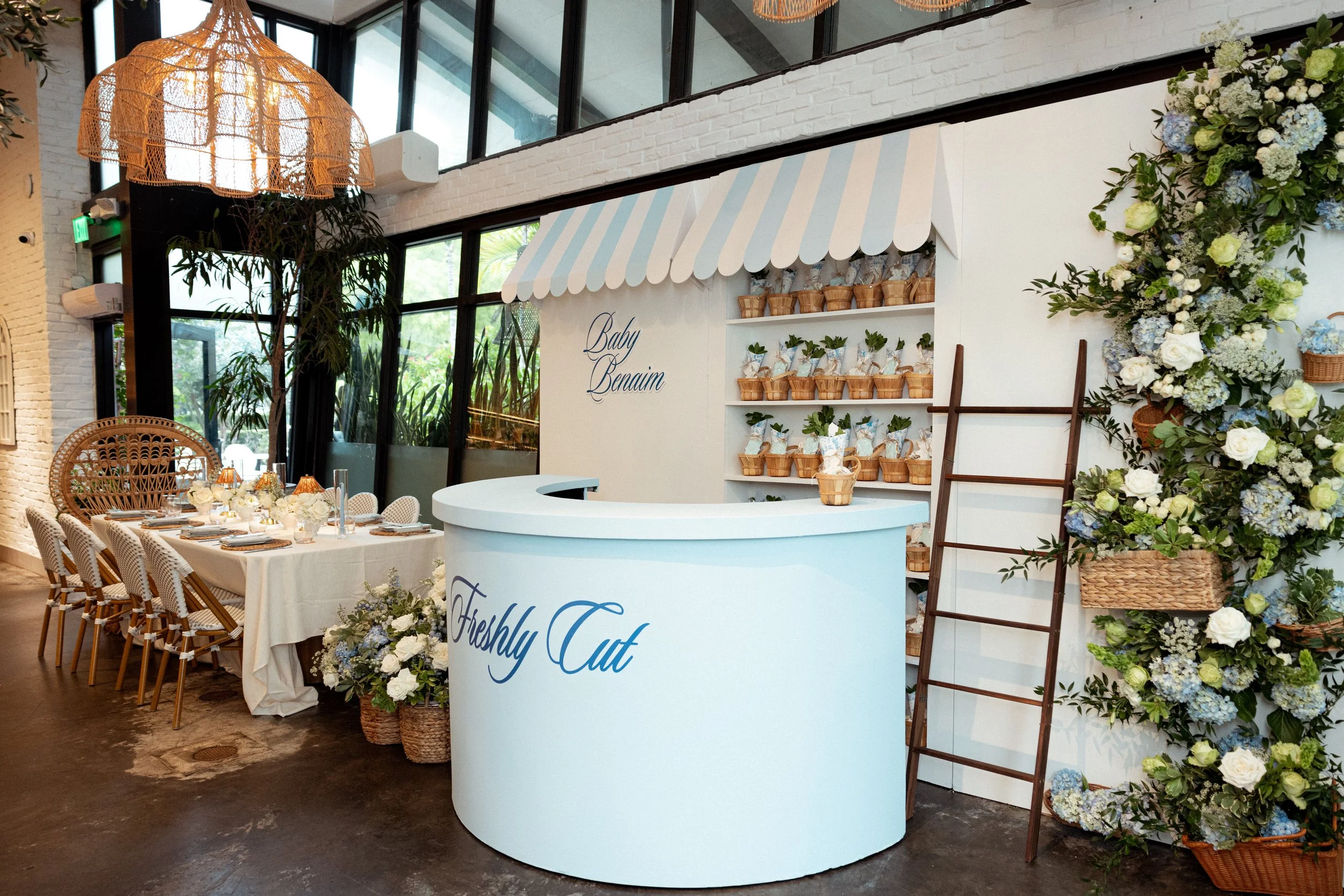 Interior of a decorated space with a table set for an event, featuring floral arrangements and a white counter with blue script reading 'Freshly Cat', in front of a floral display and a white striped canopy.
