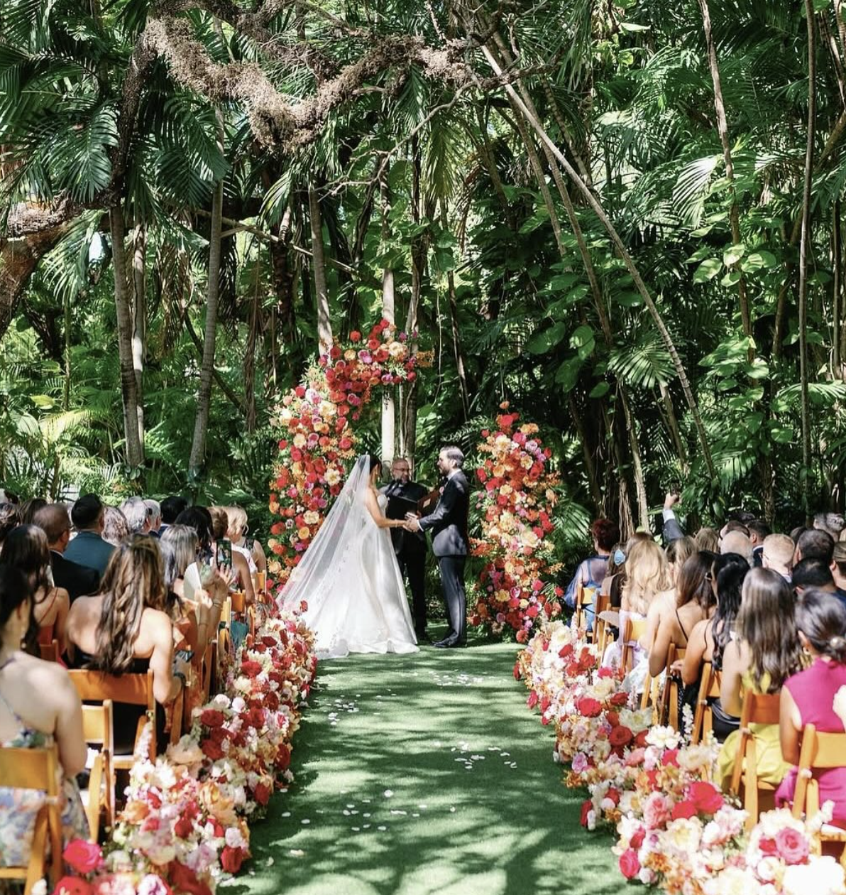 A wedding ceremony taking place outdoors in a lush jungle setting, with a bride and groom exchanging vows at an altar decorated with vibrant pink and orange flowers, surrounded by seated guests and tall green trees.