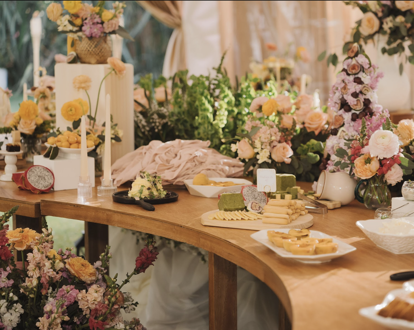 A dessert and cheese display on a wooden table decorated with an abundance of pink, purple, and cream flowers and greenery.