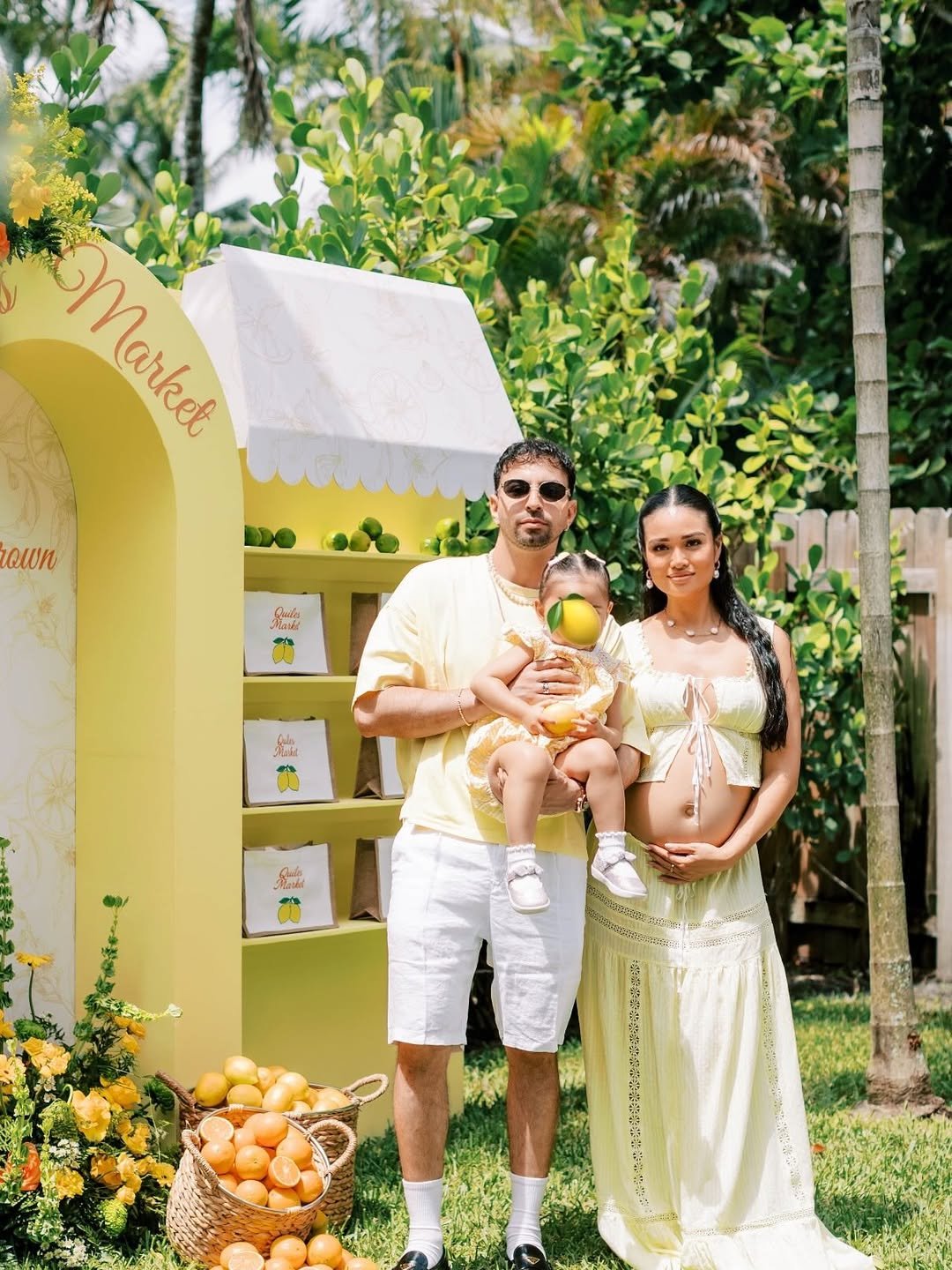 Family standing outdoors in front of lemon-themed backdrop, with basket of oranges and lemons on grass, celebrating a lemon-themed event.
