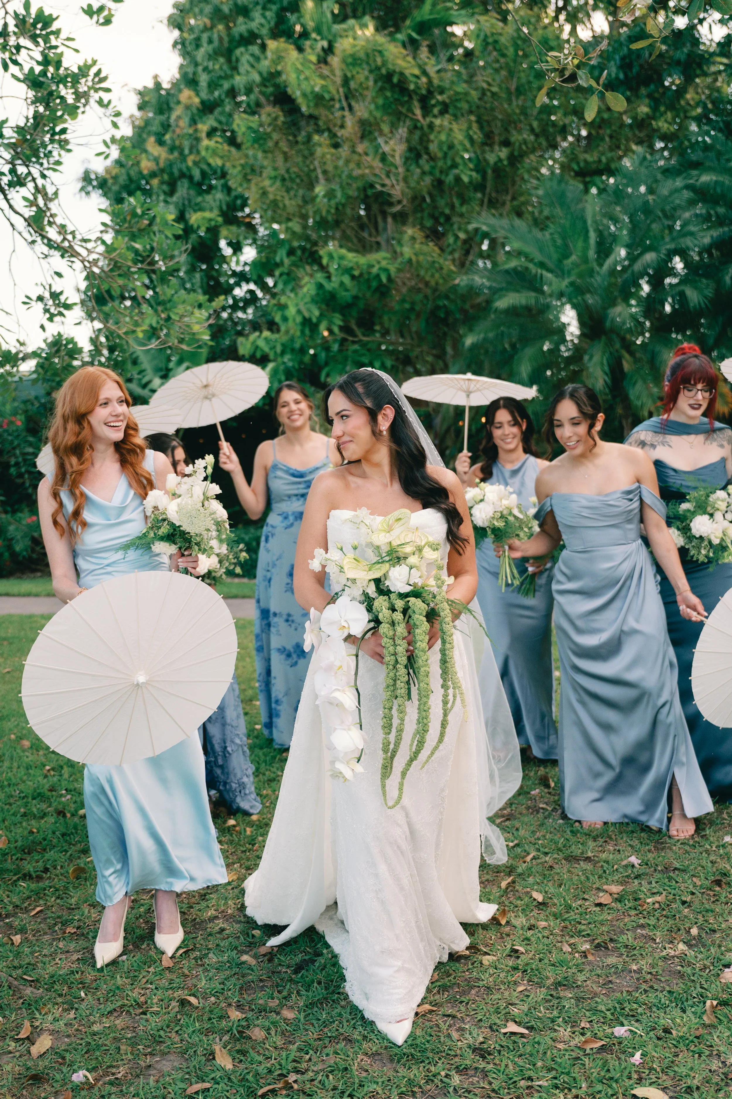 Bride in a white wedding gown holding a bouquet of white flowers walking through a garden with bridesmaids in pastel blue dresses holding white parasols and bouquets in a lush green garden.