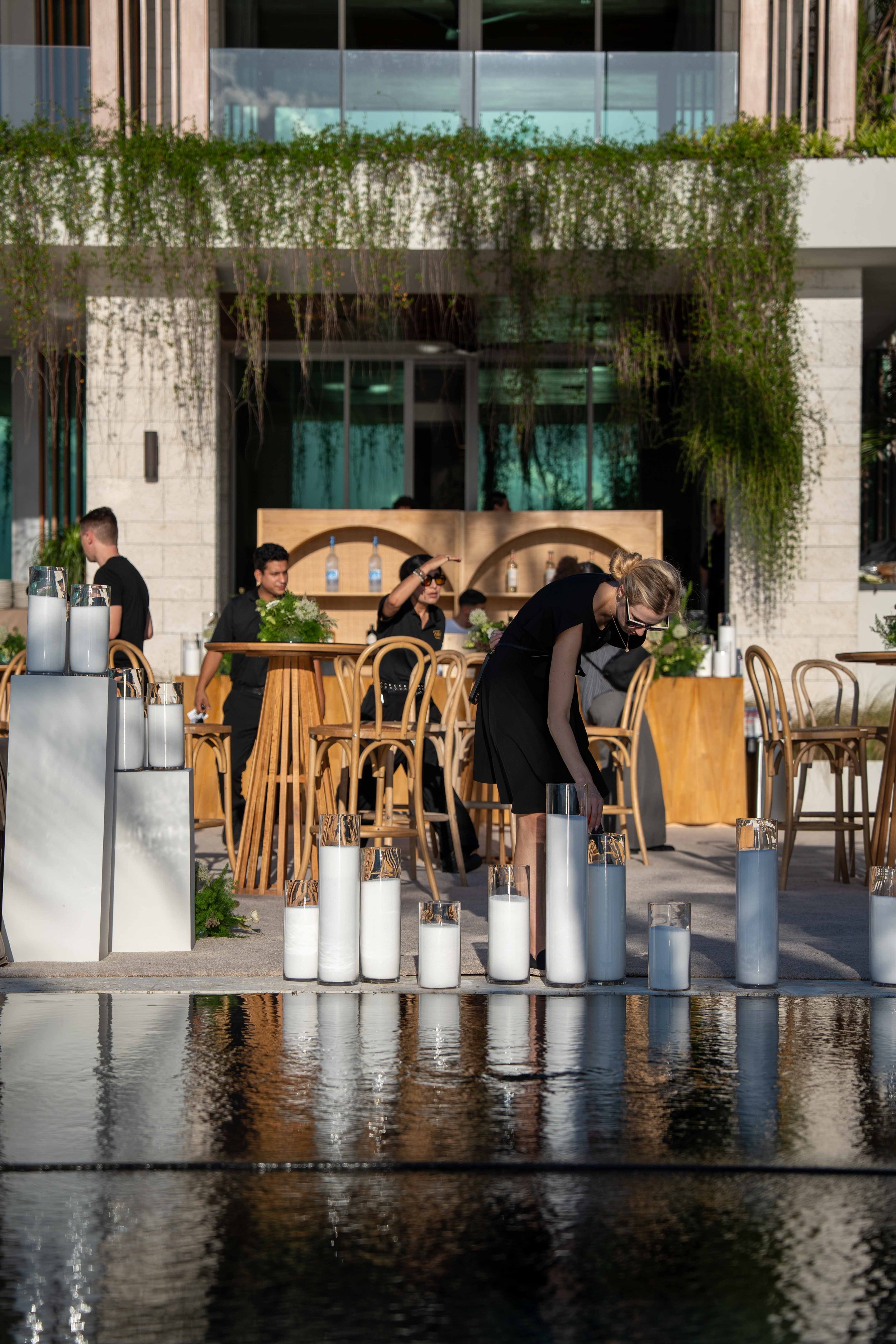 People setting up candles or lanterns on a patio near water, with a modern building featuring glass windows and greenery in the background.