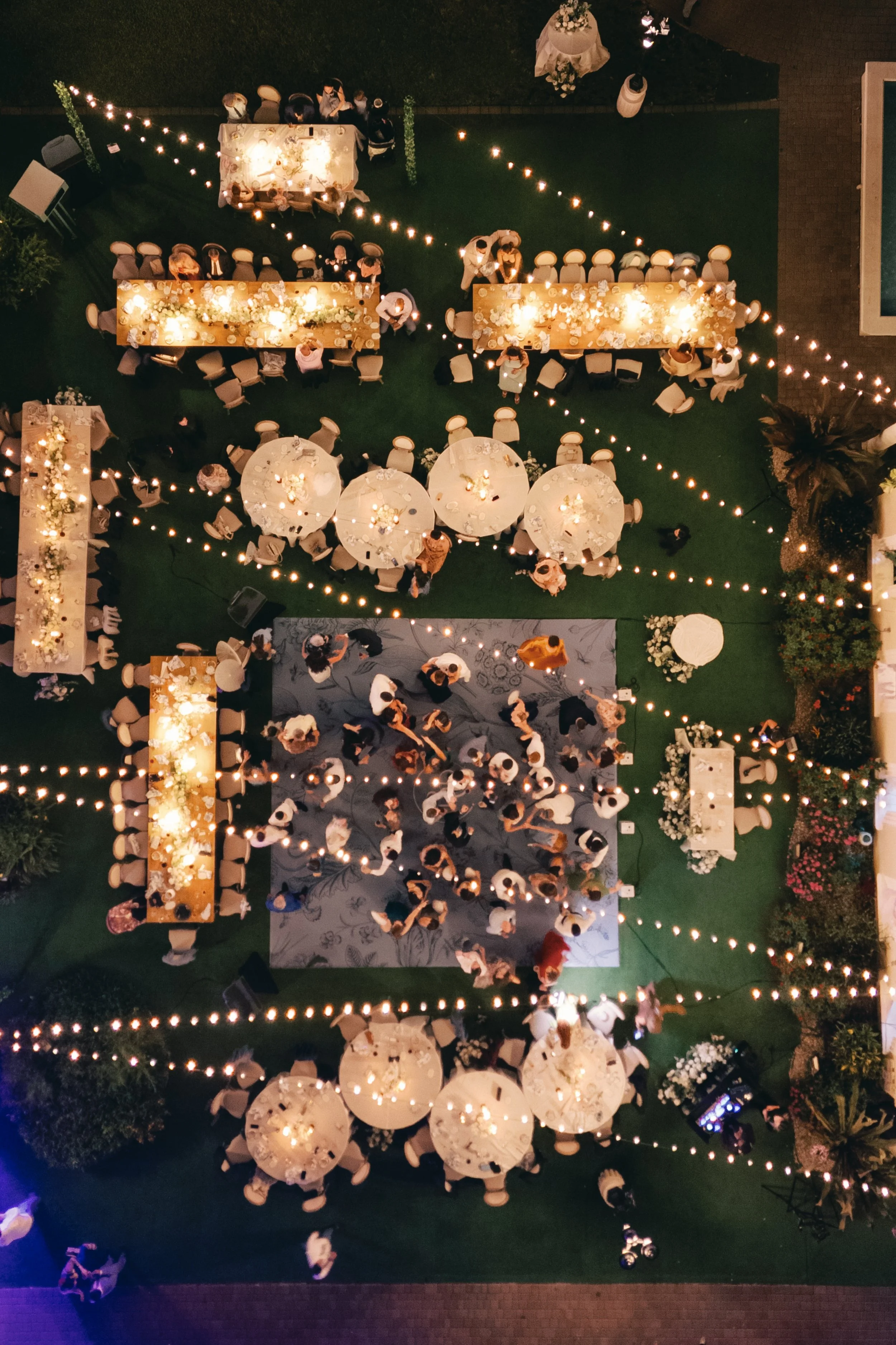 An aerial view of an outdoor evening event with round and rectangular tables decorated with flowers and candles, surrounded by chairs, and lit with string lights. Guests are mingling and dining on a grassy area with some pathways, trees, and decorati