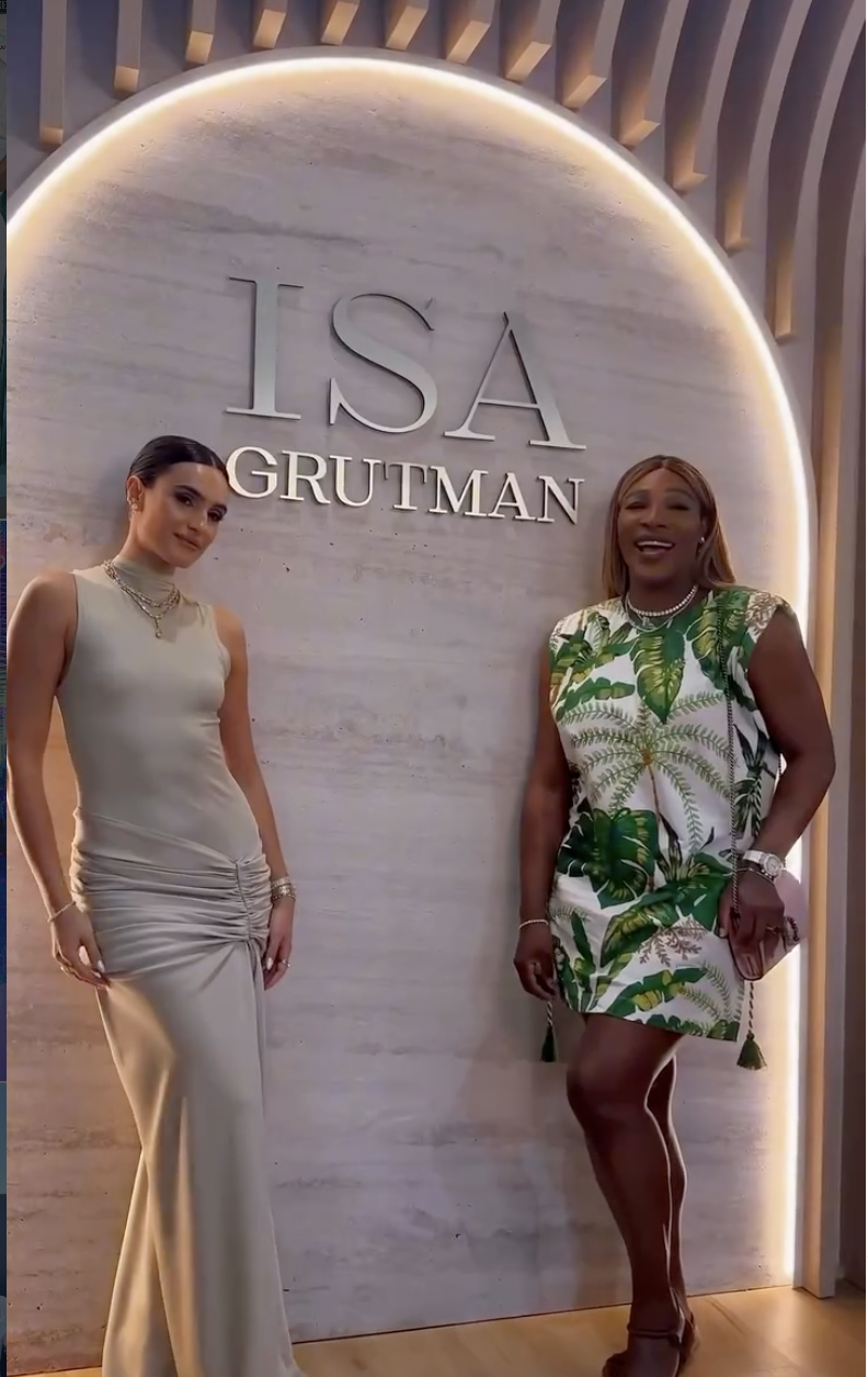 Two women standing in front of a wall with the sign 'ISA GRUTMAN'. One woman is wearing a light-colored sleeveless dress with jewelry, and the other woman is wearing a green and white tropical print dress, holding a clutch, and smiling.
