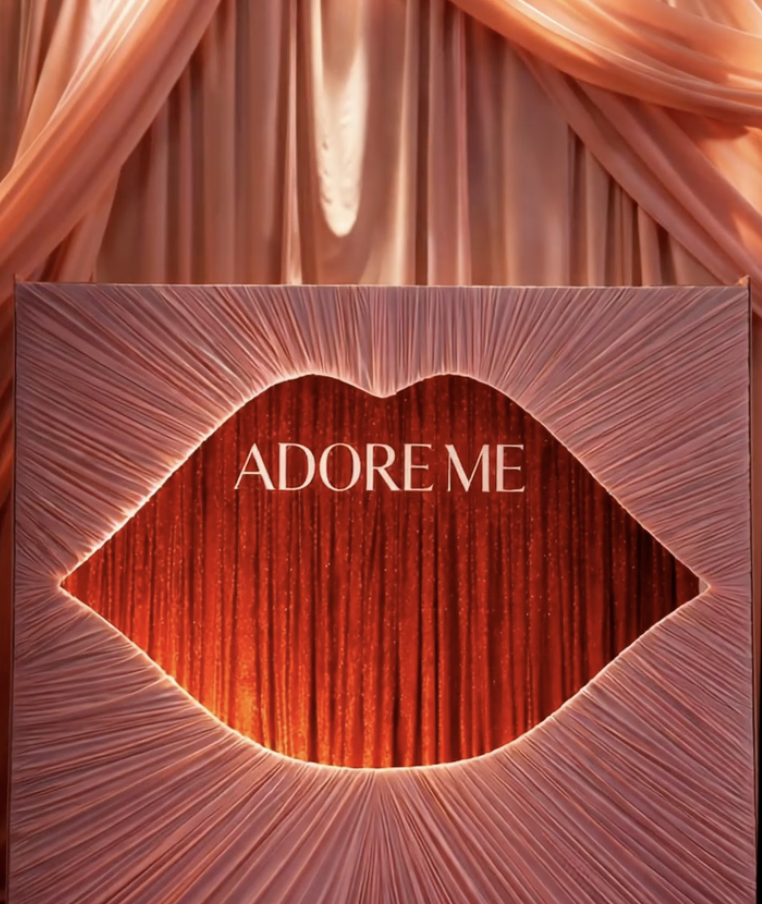 A decorative sign shaped like lips with the text "Adore Me" inside, surrounded by pink and peach drapery.