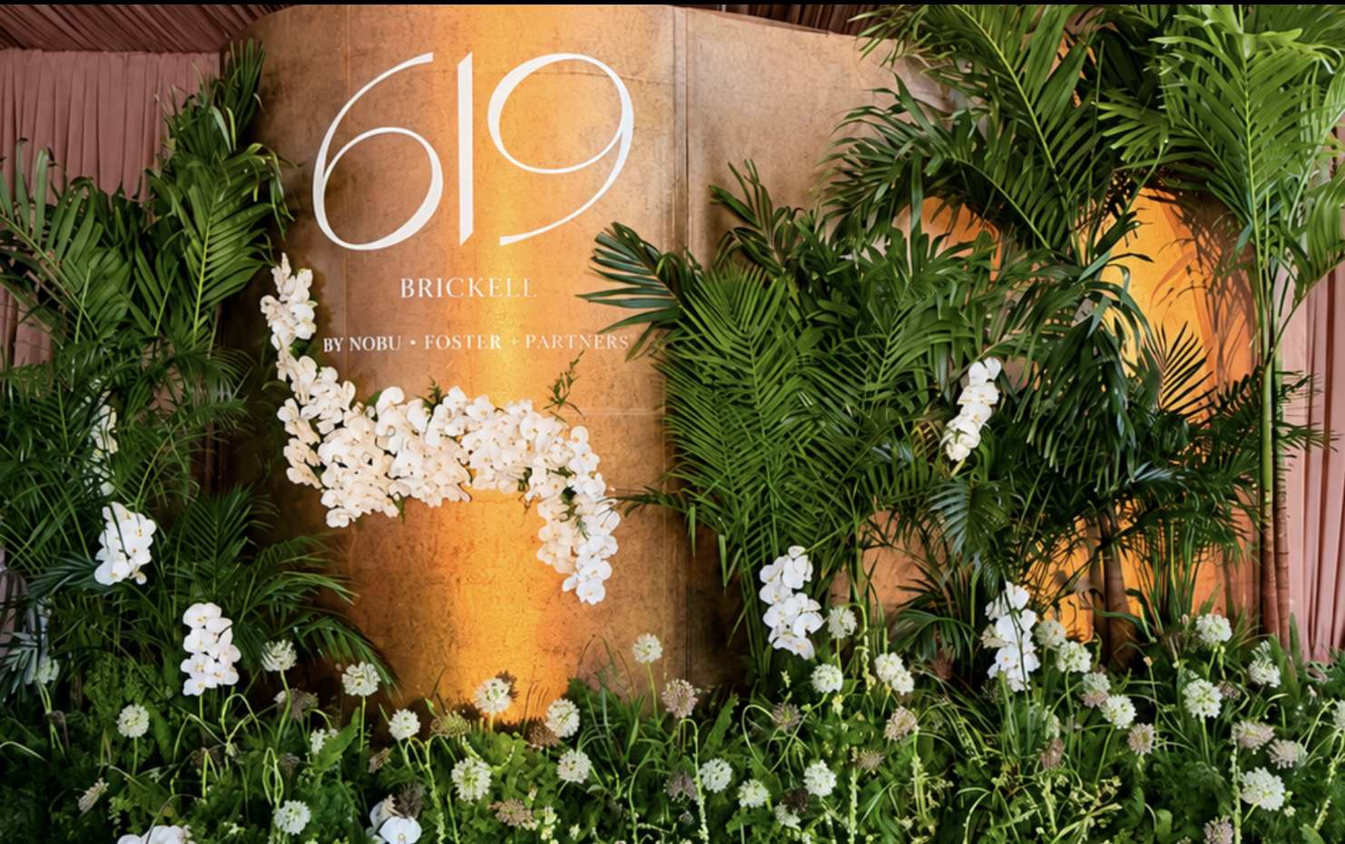 A decorative display of lush green plants and white flowers surrounding a large number 619 and the word 'Brickell' on a background with warm orange lighting.