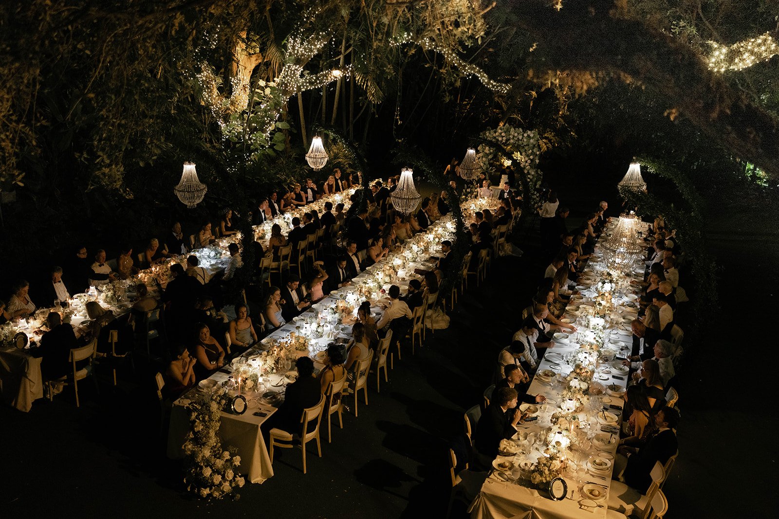 Long formal dinner table set with white tablecloths, floral centerpieces, candles, and elegant chandeliers hanging from a lush, dark green outdoor space filled with trees and hanging lights during a nighttime event.
