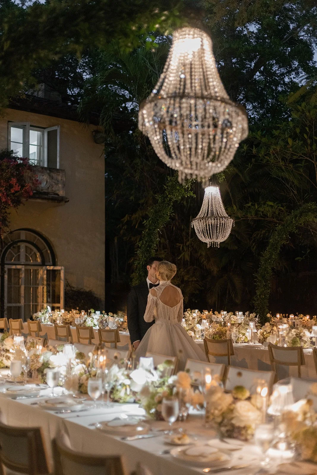 A bride and groom kissing at a wedding reception outdoor dinner setup with chandeliers hanging above, tables decorated with white flowers, candles, and elegant tableware, surrounded by trees and a building with open windows.