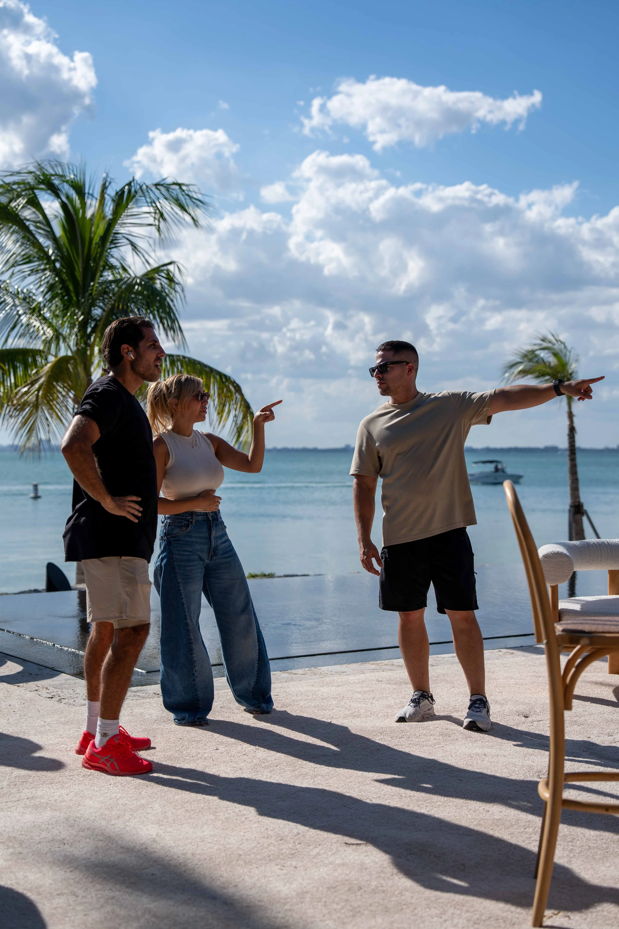 Three people stand outdoors near the water with palm trees and a boat in the background, engaging in a conversation. One man is pointing to the right while another woman gestures with her finger. The third man listens, wearing sunglasses. The scene is sunny with scattered clouds.