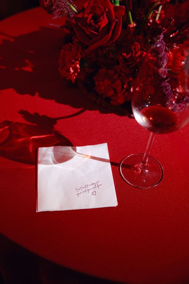 A table with a red floral centerpiece, a glass of white wine, and a white napkin with handwritten text and a heart, on a red tablecloth.