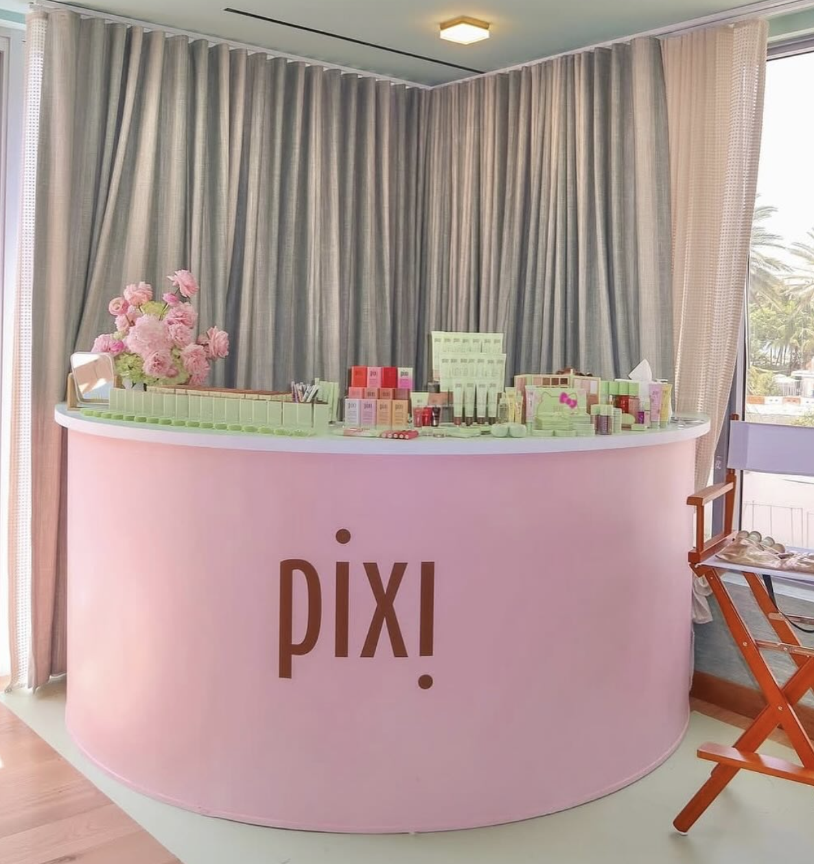 A pink curved counter with the word 'Pixi' written on it, displays various skincare and makeup products, with a bouquet of pink flowers on the left side, set against curtains with a window showing outdoor palm trees.