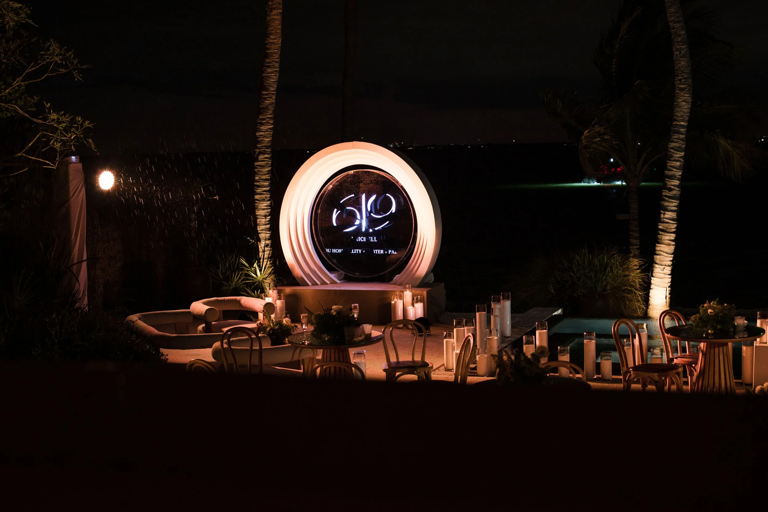 Nighttime outdoor event setup with a large illuminated circular sign, palm trees, tables with candles, and chairs near a pool, with distant city lights.