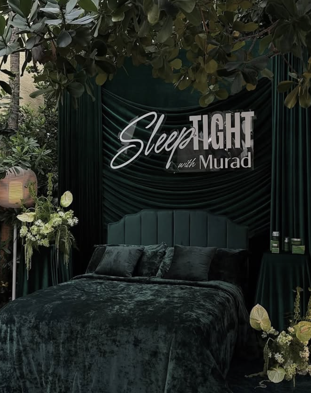 A luxurious bedroom set with dark green velvet bed and pillows, surrounded by dark green drapes and lush greenery. A sign reading 'Sleep Tight with Murad' is displayed on the backdrop.