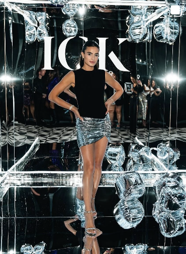 A woman standing in front of a mirror at a fashion event, wearing a black sleeveless top and a metallic silver mini skirt, with her hands on her hips. The background has reflective surfaces and large ice cube decorations, with the letters ICK visible