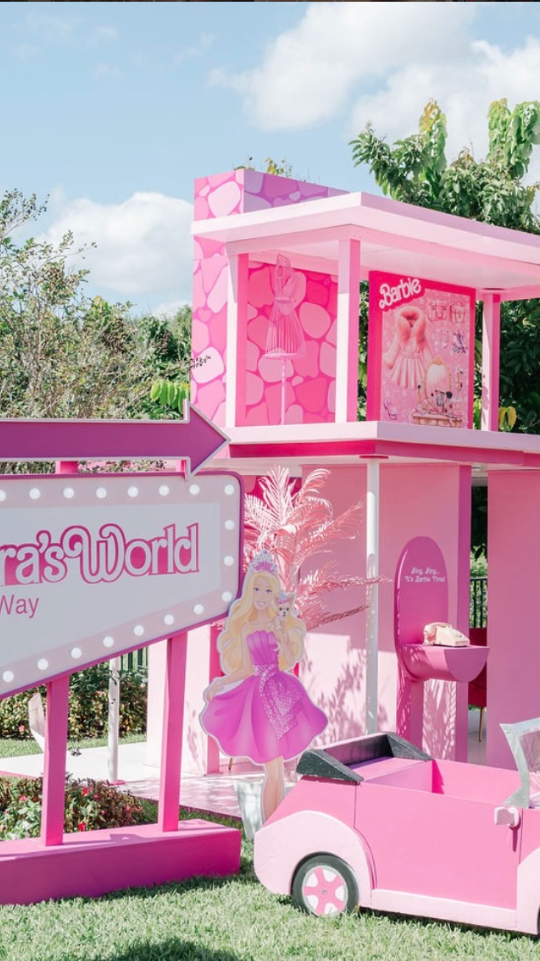 A pink themed Barbie Dreamhouse with a sign reading 'Barbie's World' and a cardboard cutout of Barbie in a pink dress. The house has multiple levels and a carport with a pink toy car.