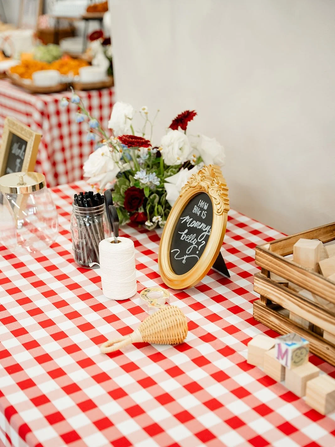 A table with a red and white checkered tablecloth, decorated with a small chalkboard sign in an ornate gold frame that says 'How big is mommy belly?'. There is a floral centerpiece with white and red flowers, a jar of black pens, a roll of white thre