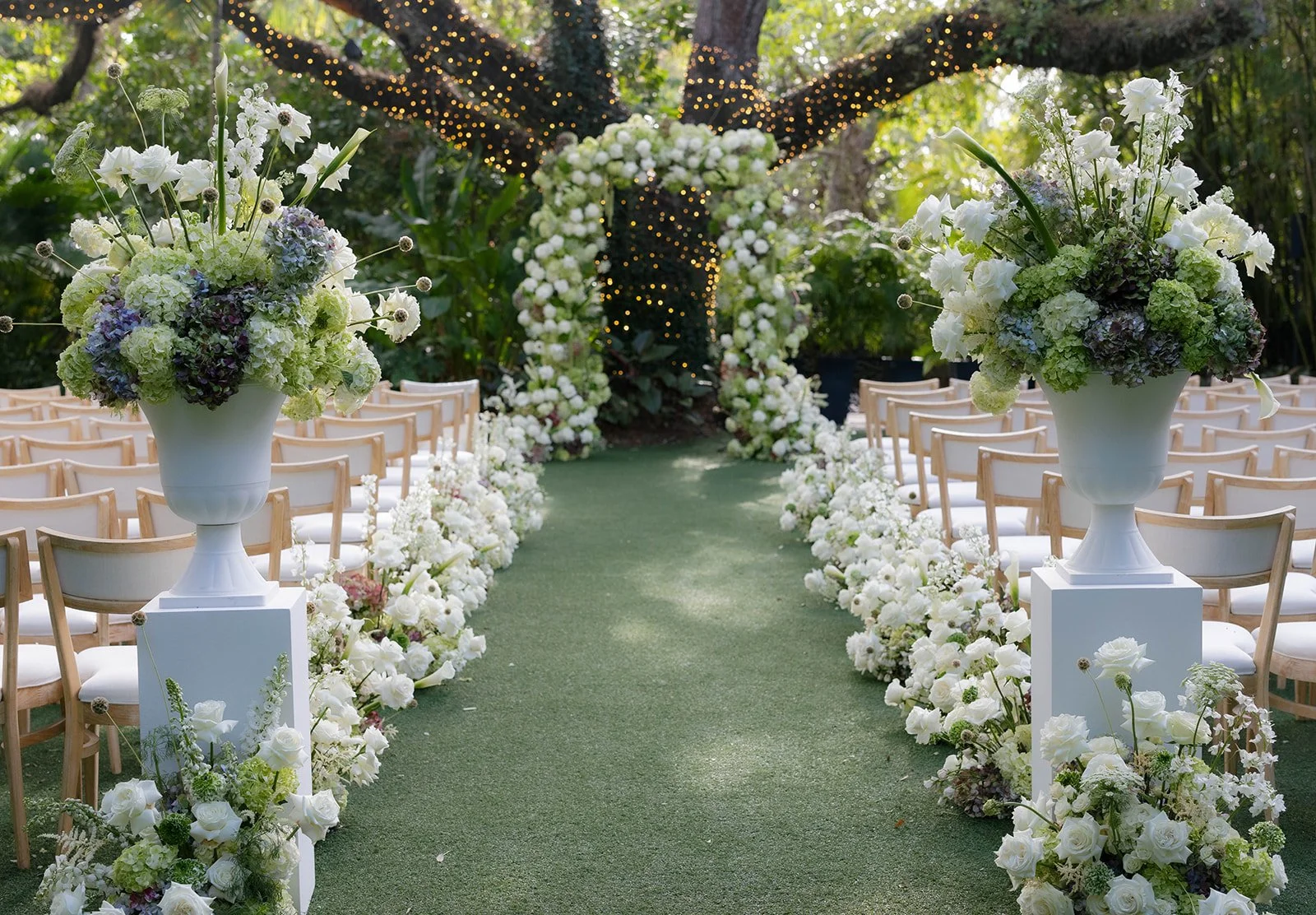 Outdoor wedding ceremony setup with white chairs on either side of a grassy aisle, lined with white floral arrangements on pedestals and arches covered in white flowers. String lights are hanging from trees in the background.