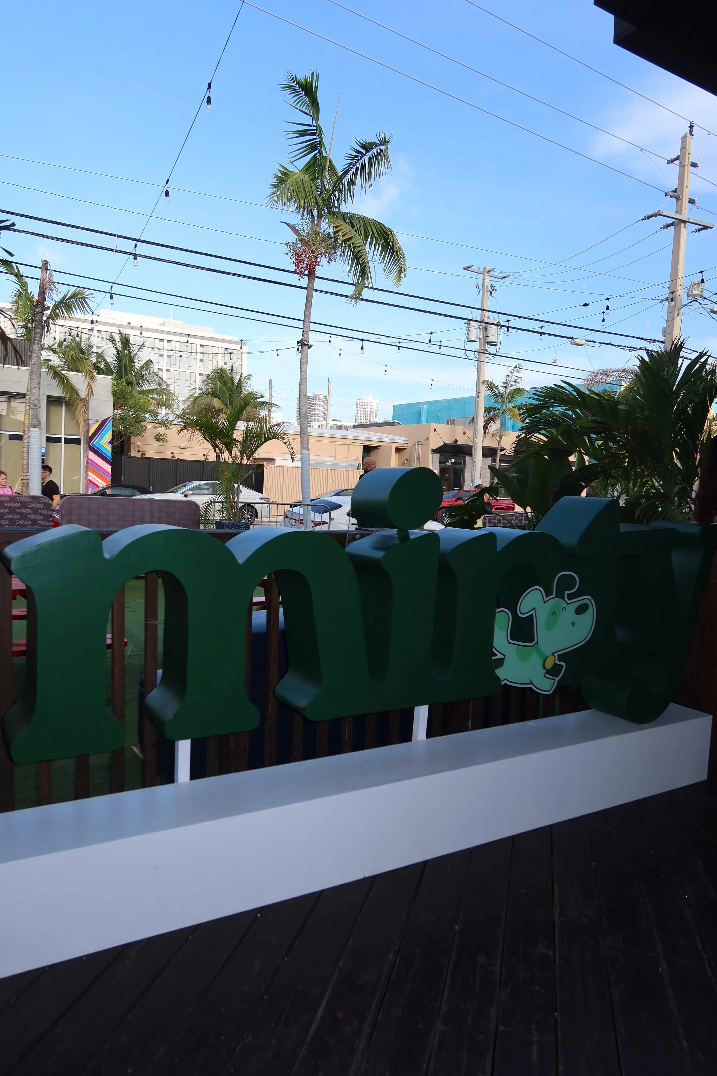 Outdoor scene with palm trees, string lights, parked cars, and a building in the background, viewed from inside a restaurant or cafe with seating and decorative green cutouts in the foreground.