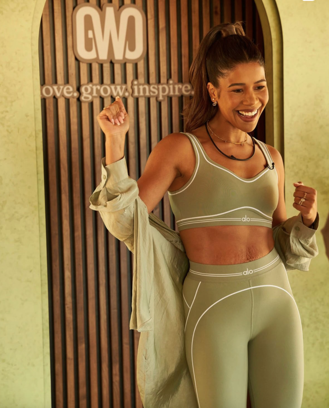 A woman smiling and flexing her arms in front of a wooden wall with the words "love. grow. inspire" and the initials "G W" behind her. She is wearing a matching green sports bra and leggings from Alo, with a beige jacket off her shoulders.