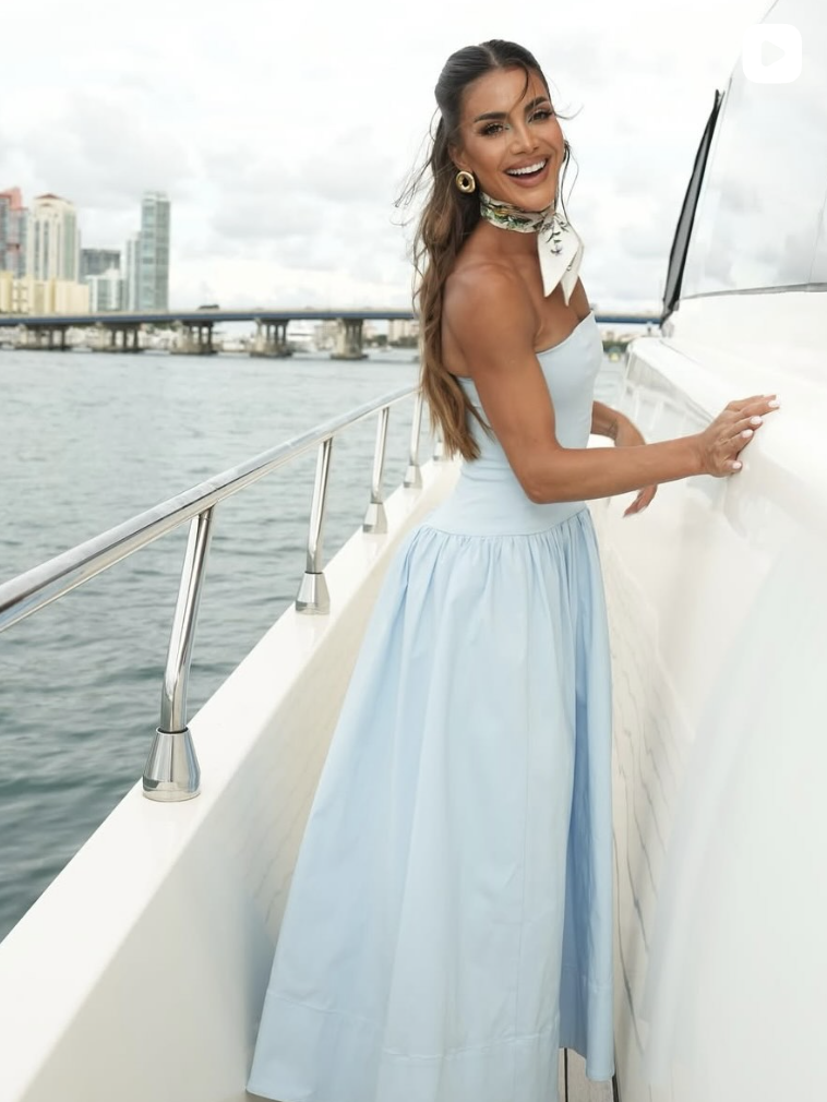 A woman in a light blue gown smiling and standing on a boat with city buildings and a bridge in the background.