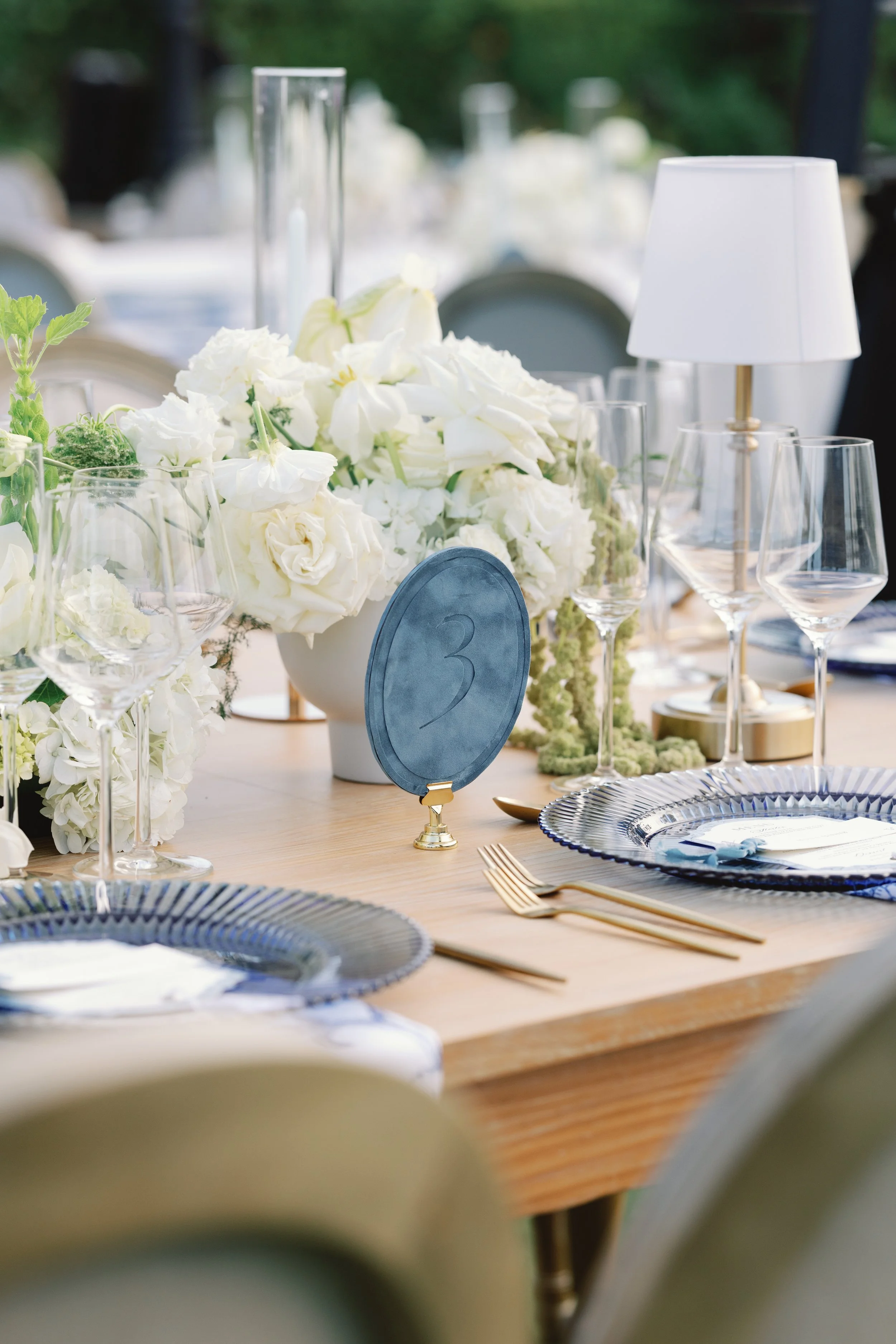 Elegant outdoor dining table decorated with white floral centerpiece, glassware, golden cutlery, blue and white plates, and a table number card, set in a lush garden setting.