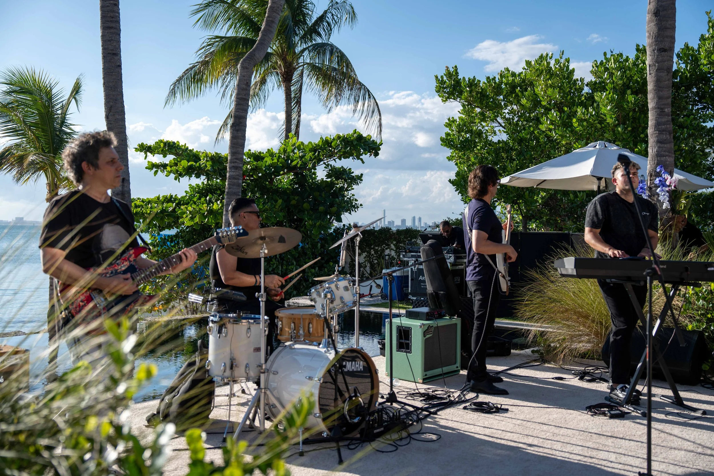 Musicians performing outdoors near a body of water, with palm trees and green bushes in the background, under a partly cloudy sky, in a tropical setting.