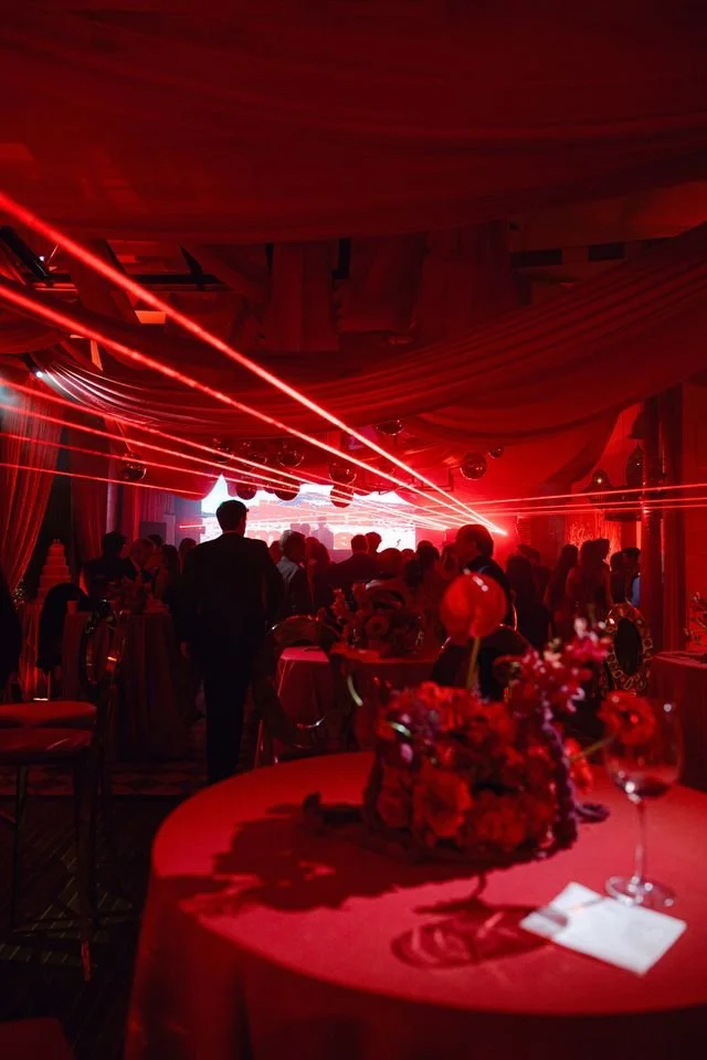 Indoor event with dim red lighting, decorated tables, and laser lights, likely a party or celebration.