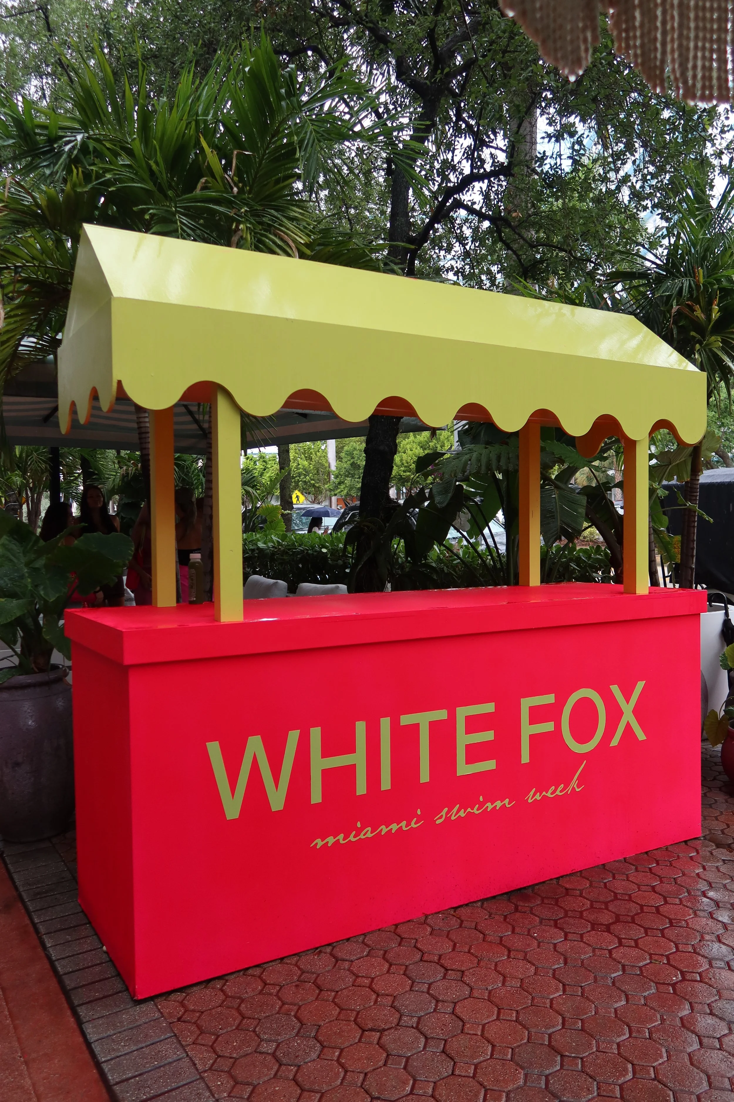 A pink and yellow stand with a yellow canopy and pink base, located outdoors among trees, with the words 'WHITE FOX' and 'miami swim week' written on the front.