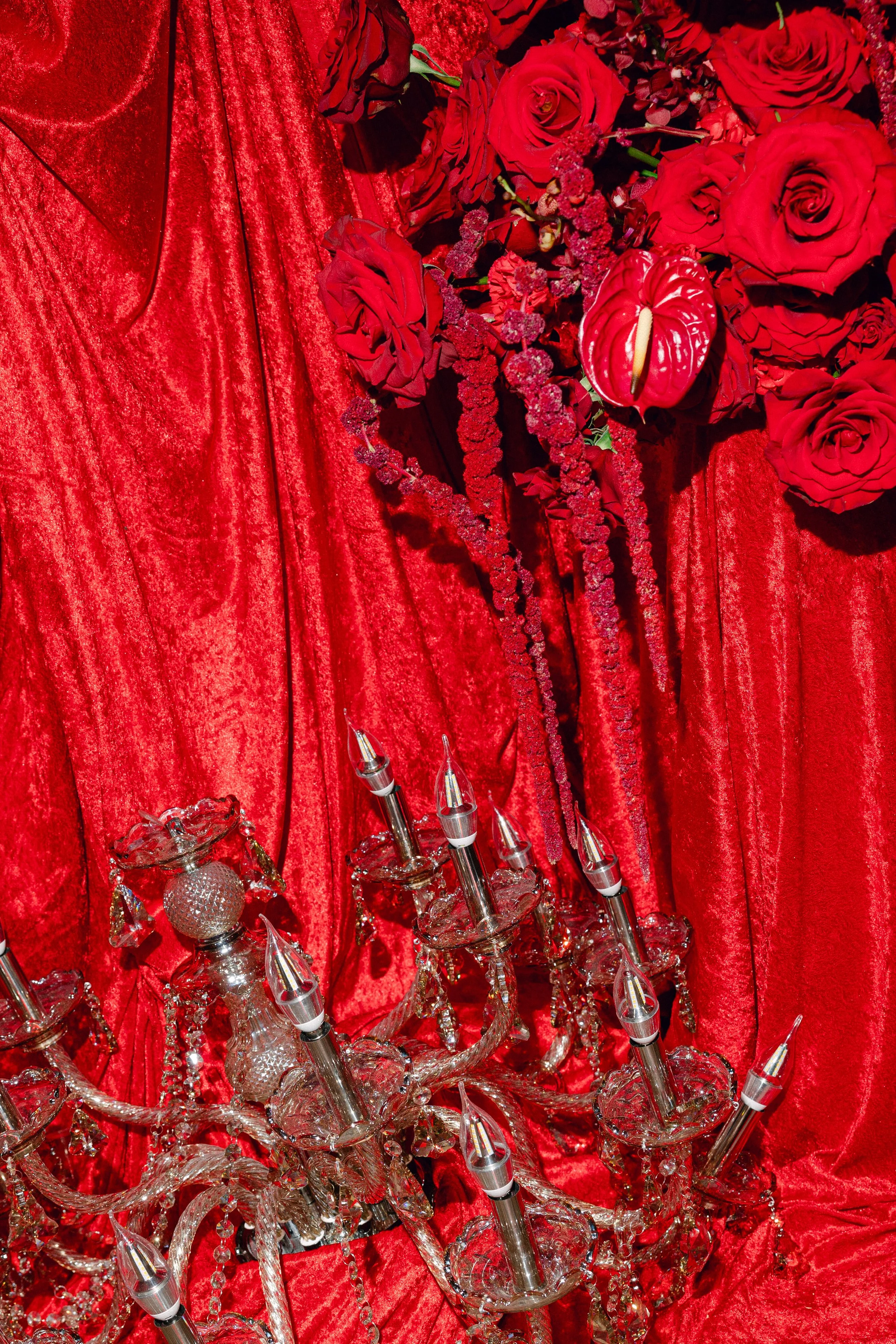 Red velvet curtain background with red roses, pink flowers, and a glass chandelier with clear, pointed crystal accents and candle-shaped light bulbs.