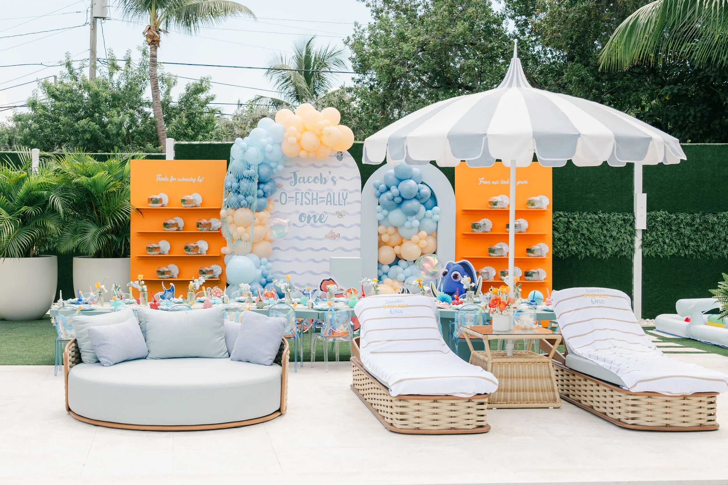 Outdoor birthday party setup with a large white umbrella, colorful balloons, and decorations themed around a mermaid or ocean. There are tables with party favors, a white couch with cushions, and lounge chairs. The background shows trees, plants, and
