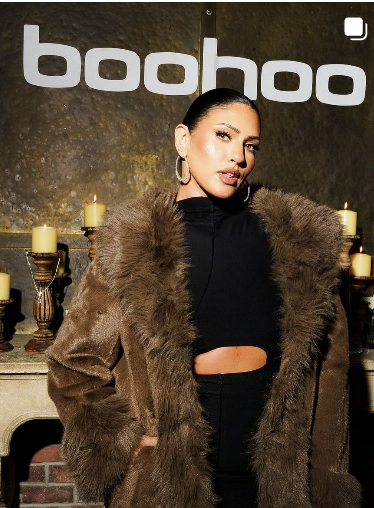 A woman in a fur coat poses in front of a dark wall with the word "bohoo" in white letters, surrounded by candles.