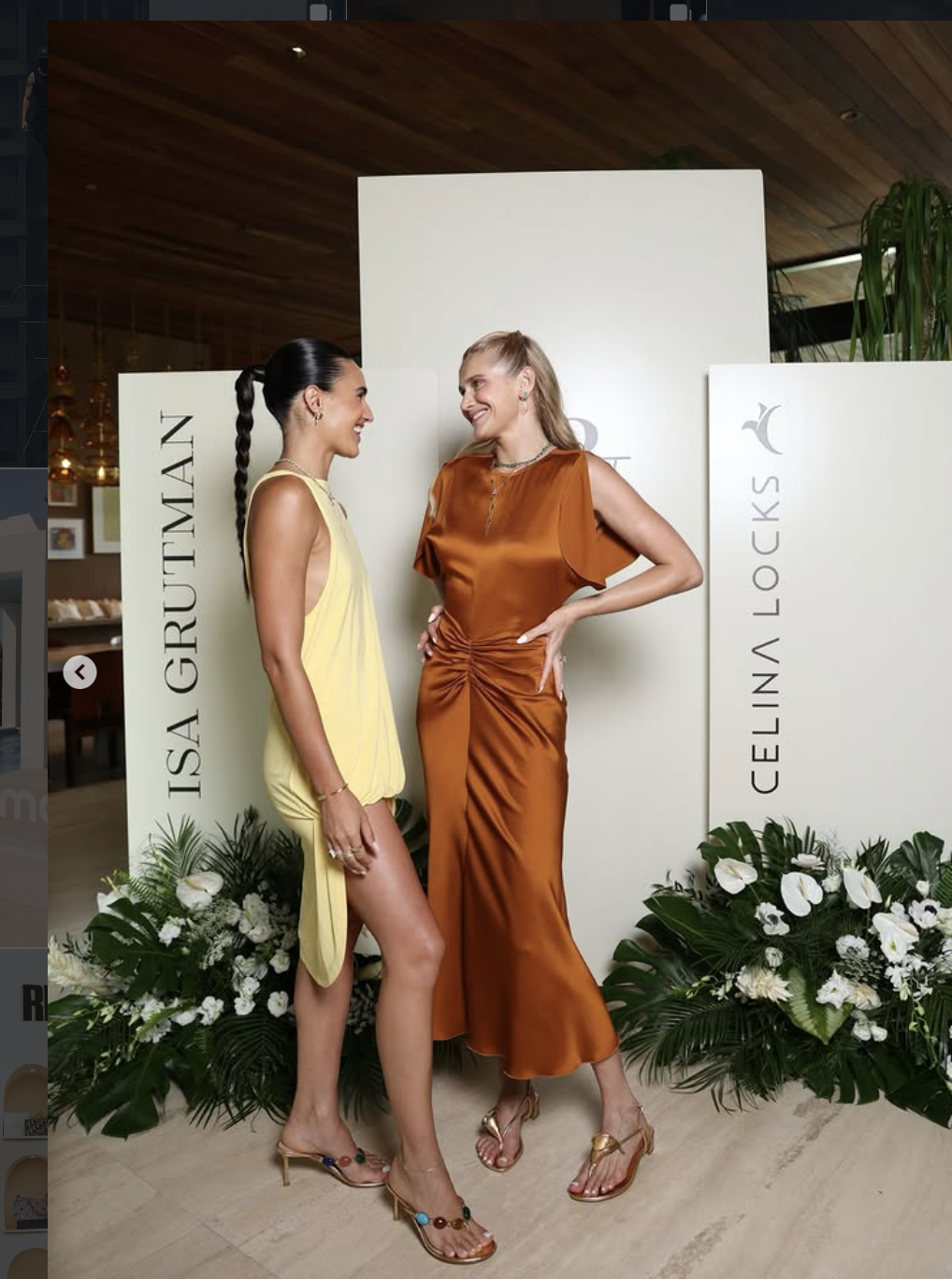Two women standing and smiling at each other in front of white panels with black text, floral arrangements at their feet, and a wooden ceiling in the background. One woman is wearing a yellow dress, and the other is in an orange dress.