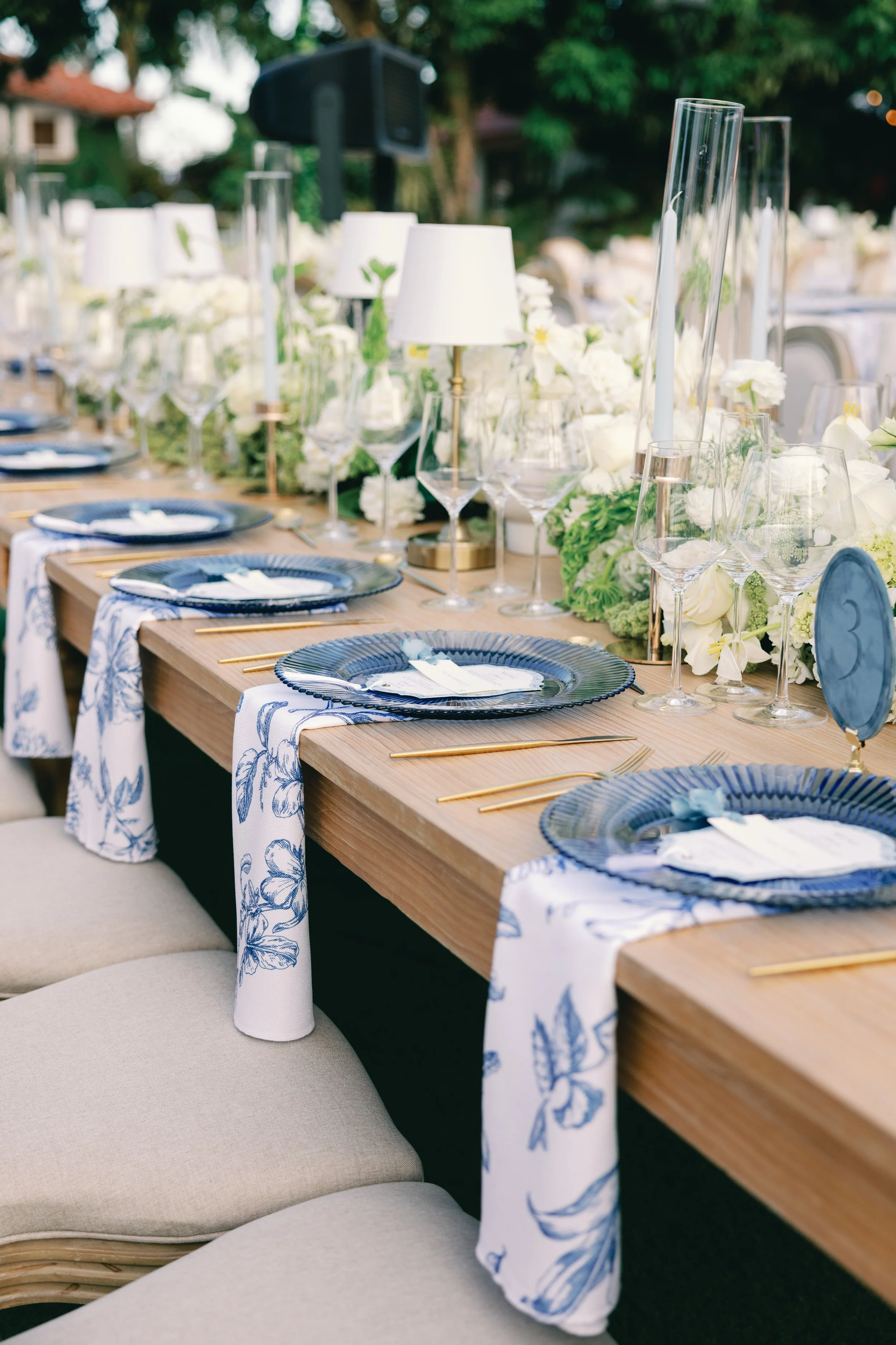 A long outdoor dining table decorated with white floral arrangements, white lamps, and napkins, set with blue plates and gold utensils, with chairs around it, in a garden setting.
