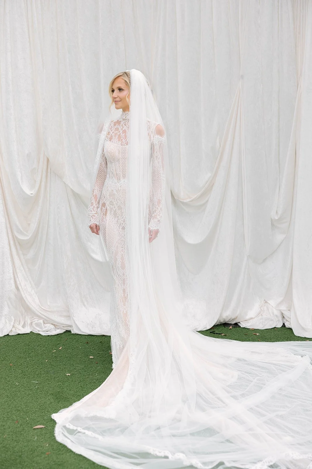 A woman in a white lace wedding dress and veil standing in front of a white curtain backdrop.