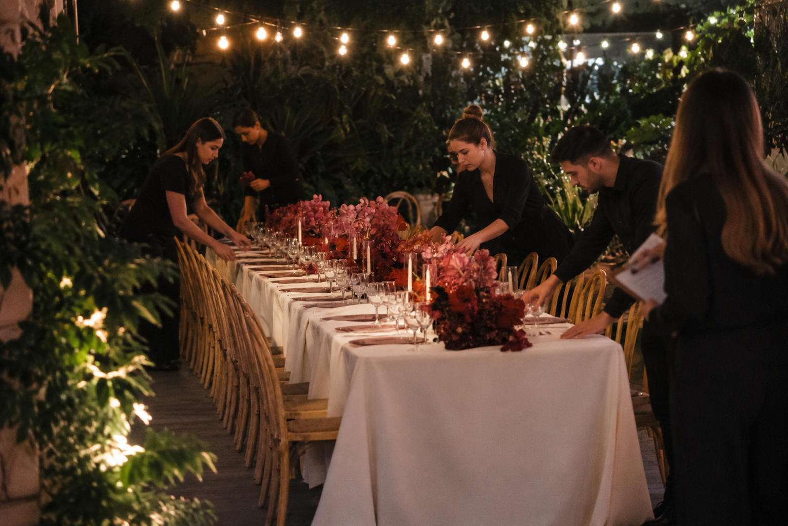 A long outdoor dining table decorated with pink and burgundy floral arrangements and candles, set for a dinner gathering at night. Several people in black attire are arranging the table, under string lights with greenery in the background.