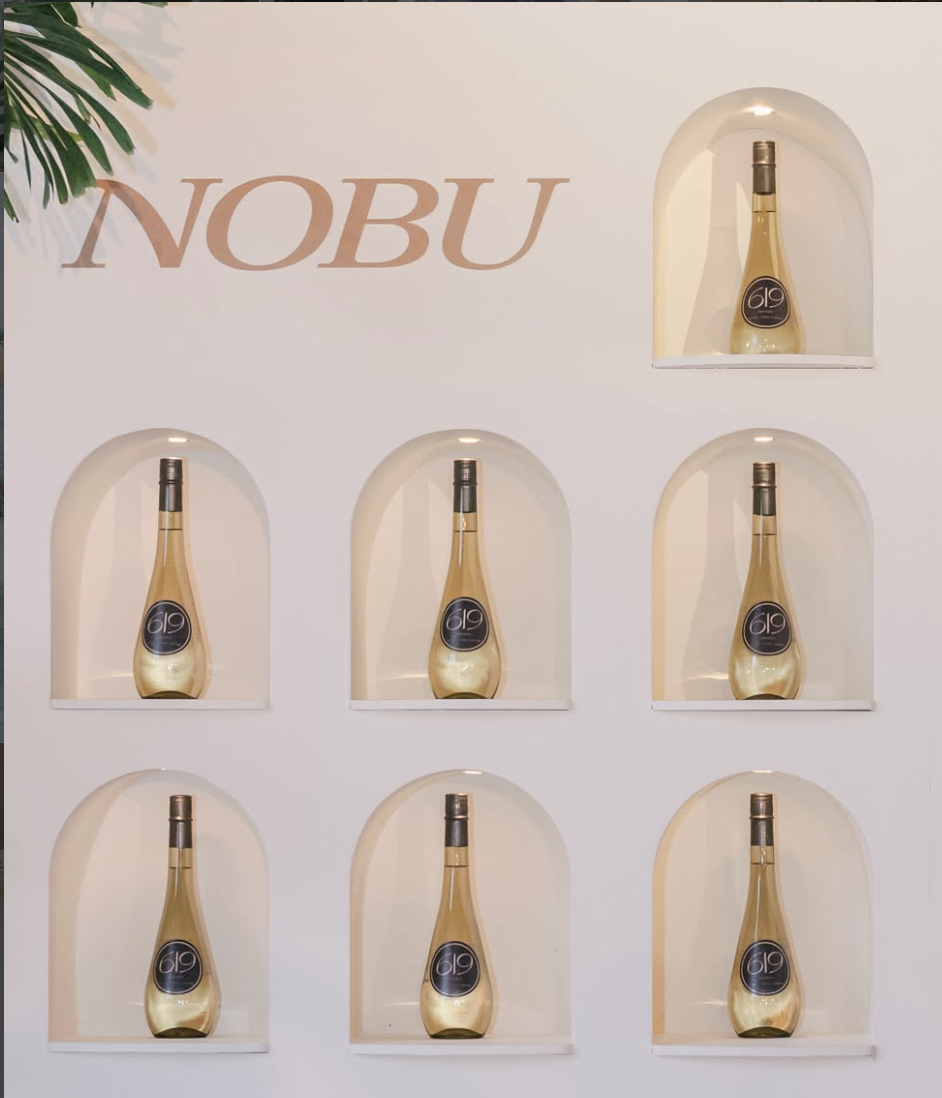 Display of six bottles of Nobu sake arranged in three rows of two on a white wall. The bottles are in individual arched niches and labeled with a black and gold sticker reading '619'. The Nobu logo is partially visible on the wall above the bottles.