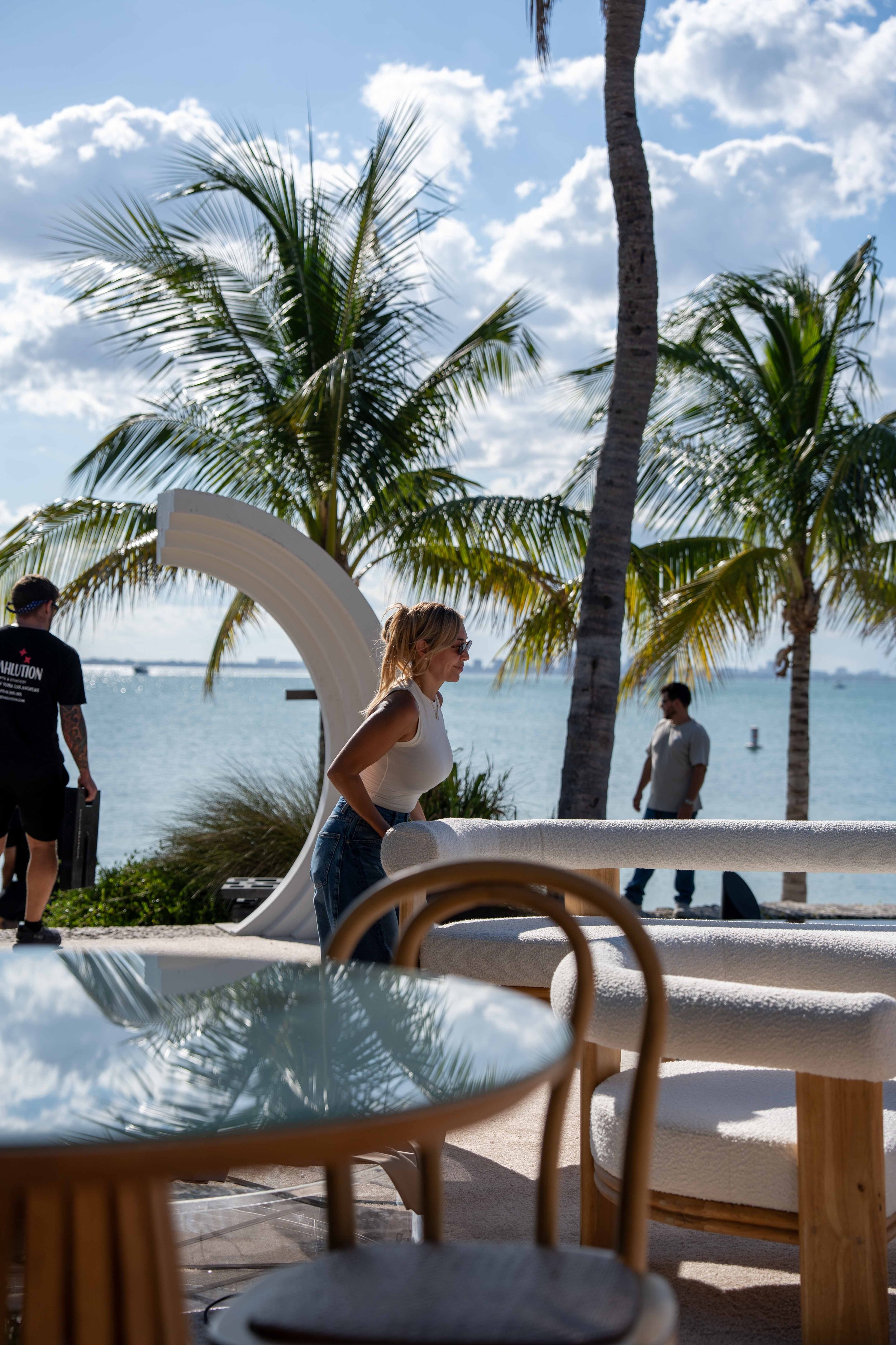A woman in a white top and jeans standing outside near the beach, with palm trees in the background and a partly cloudy sky.